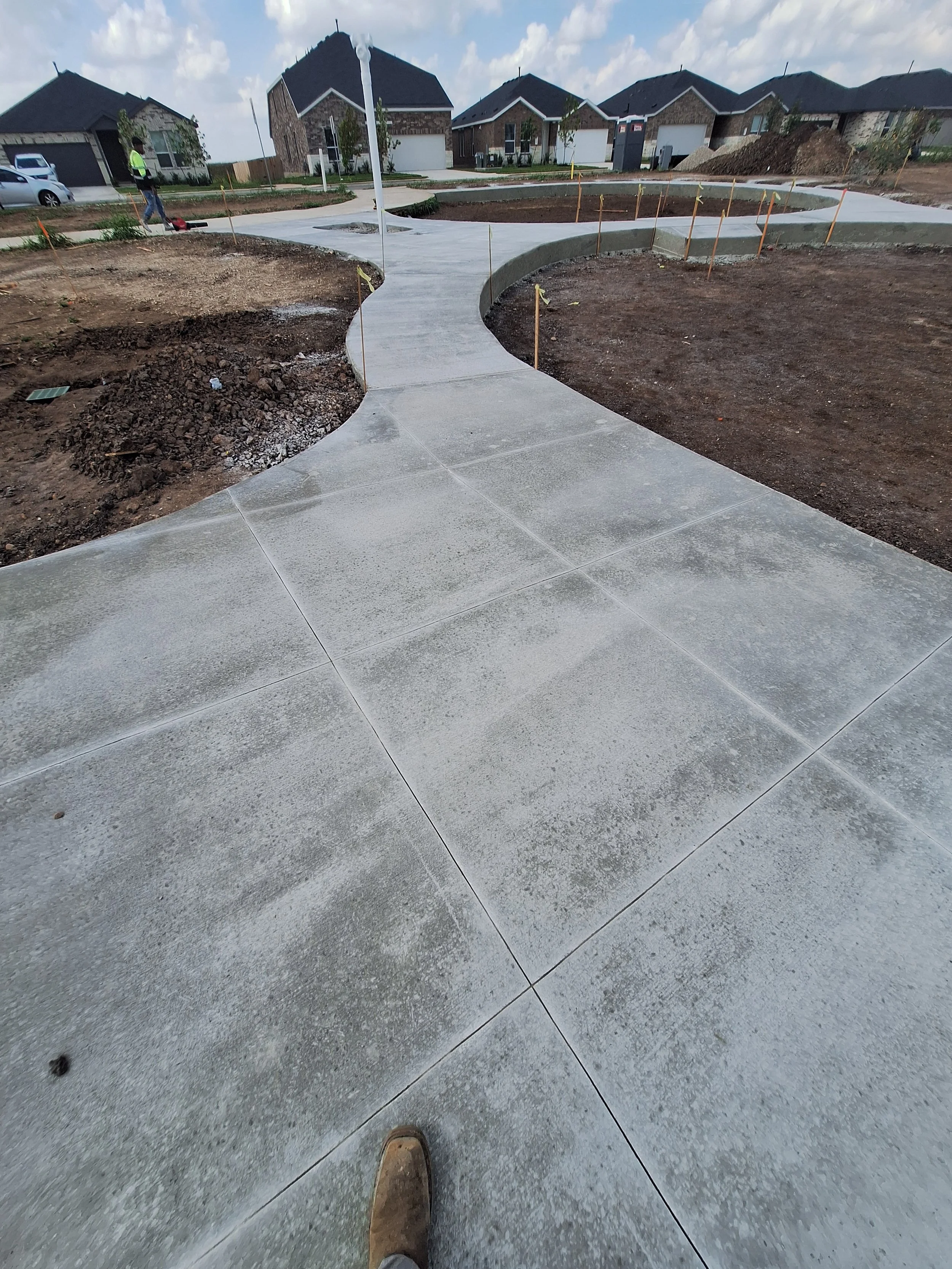 Newly poured concrete sidewalk with curves in a residential neighborhood, surrounded by bare soil and construction markers, with houses in the background.