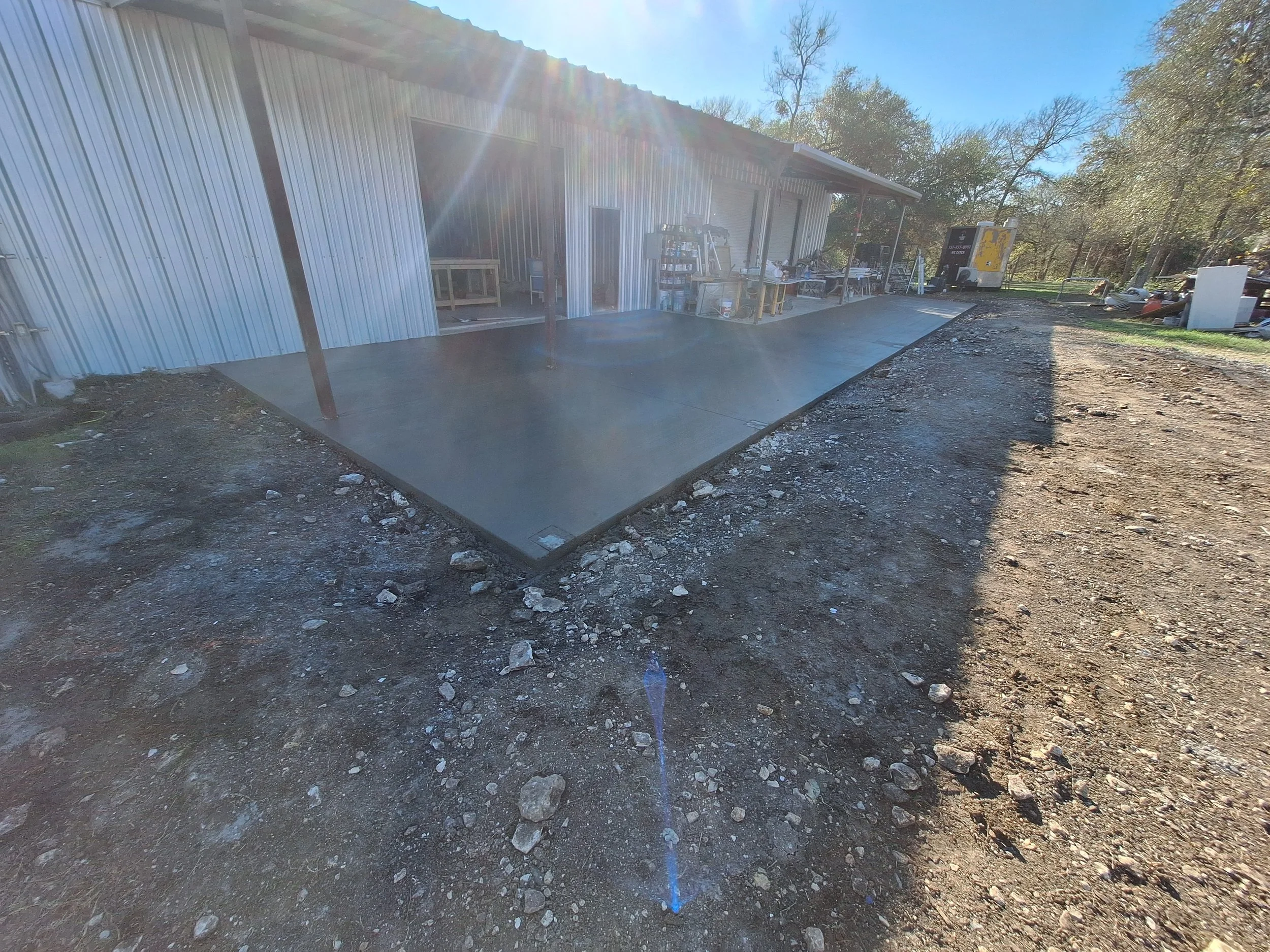 Exterior of a building with a freshly poured concrete slab and outdoor work area, with tools and equipment visible, on a sunny day with trees in the background.