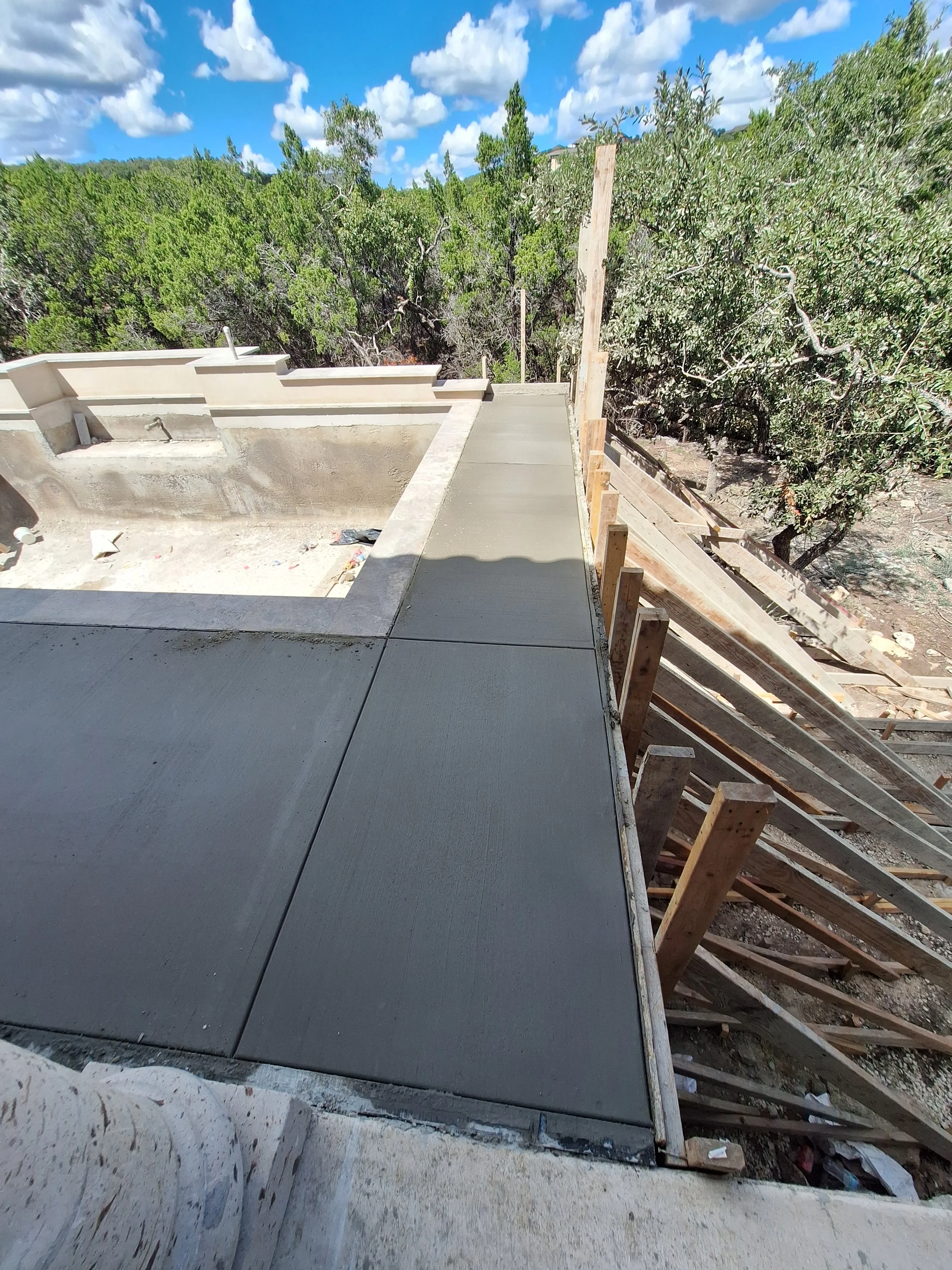 Construction site with concrete sidewalk finished, wooden supports and framing for a new structure, with trees and a partly cloudy sky in the background.
