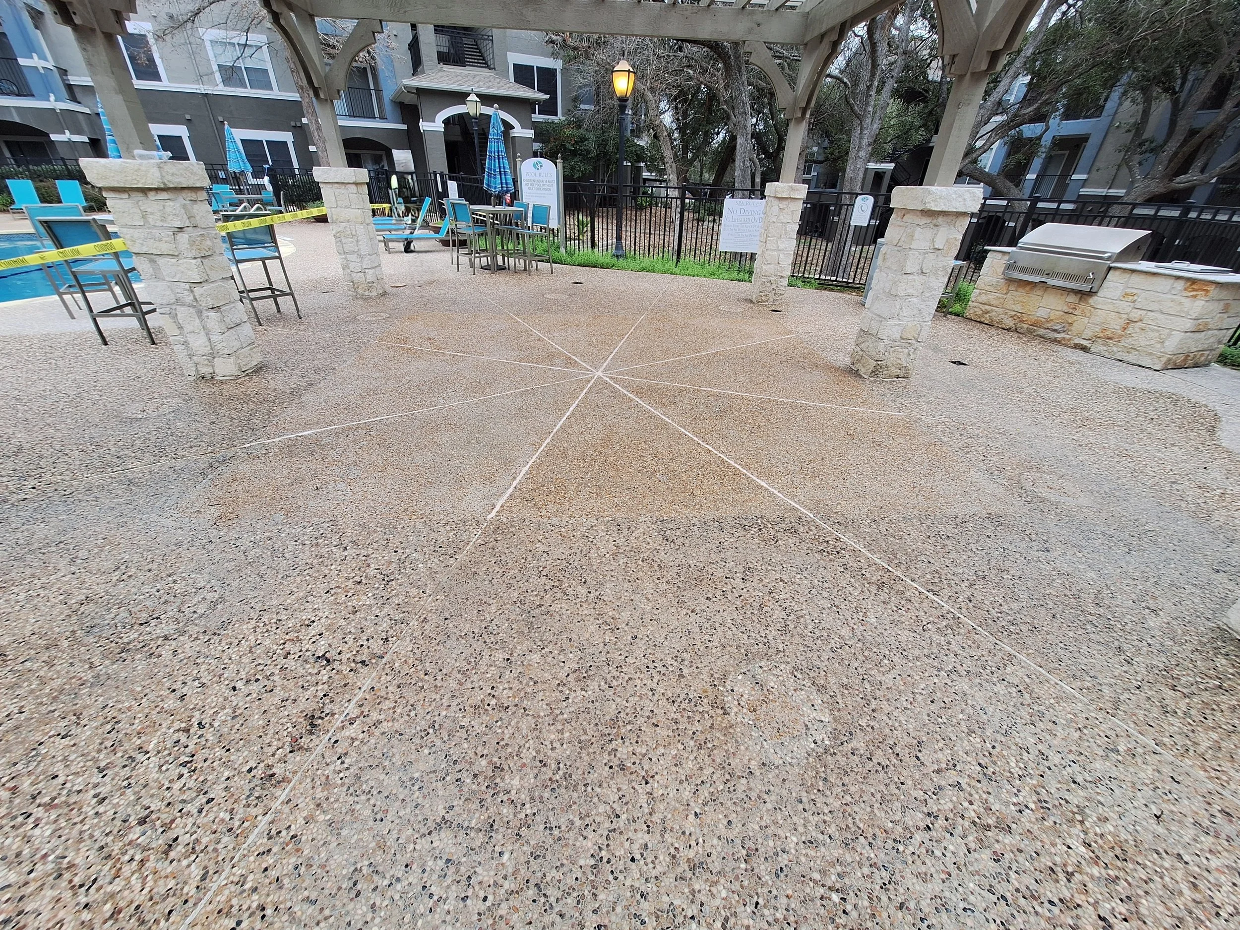 Public outdoor pool area with a canopy, patio furniture, and safety tape across the ground, surrounded by residential buildings and trees
