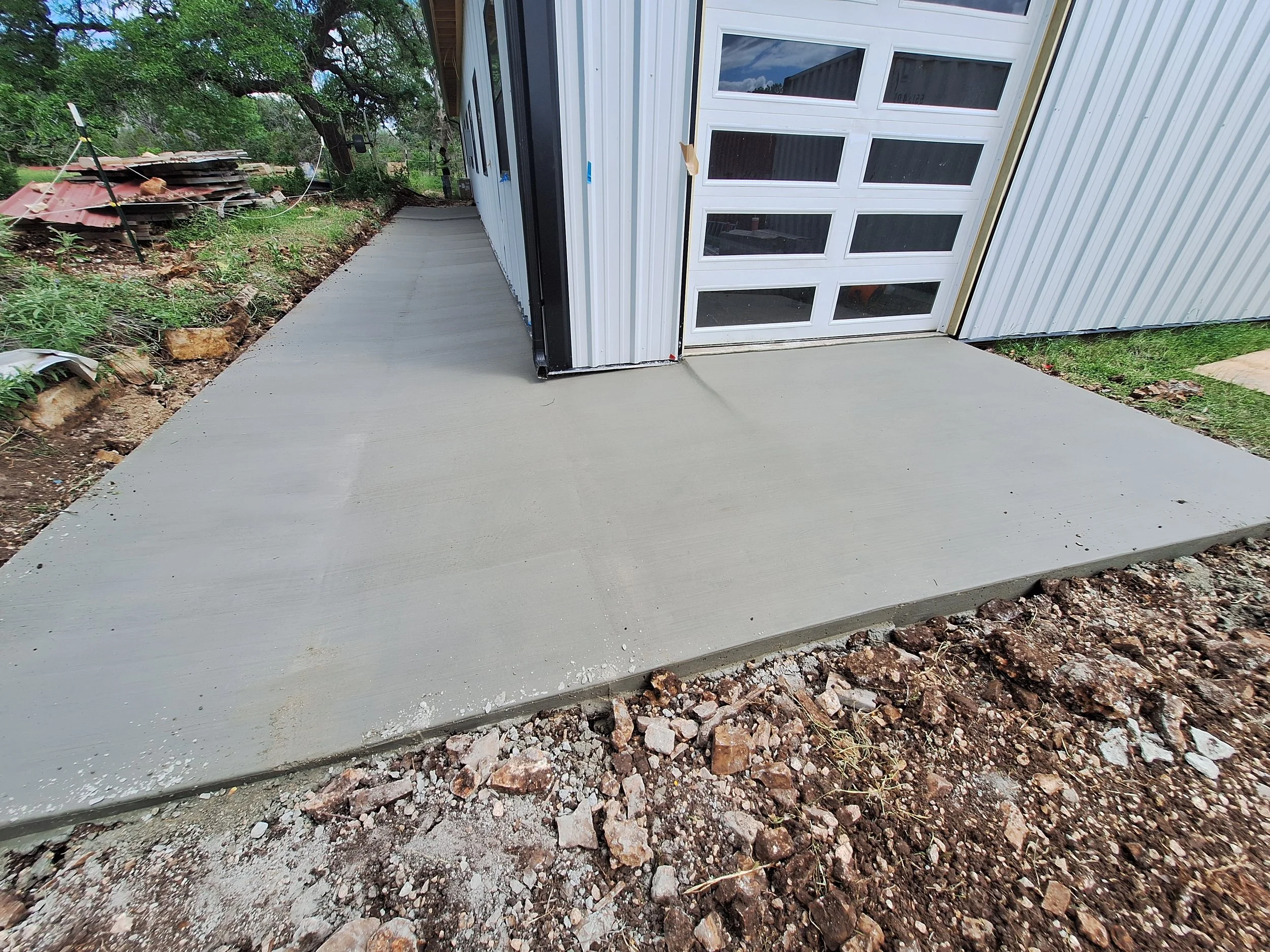 Freshly poured concrete sidewalk outside a garage with a translucent door, surrounded by a construction site with dirt and rocks.