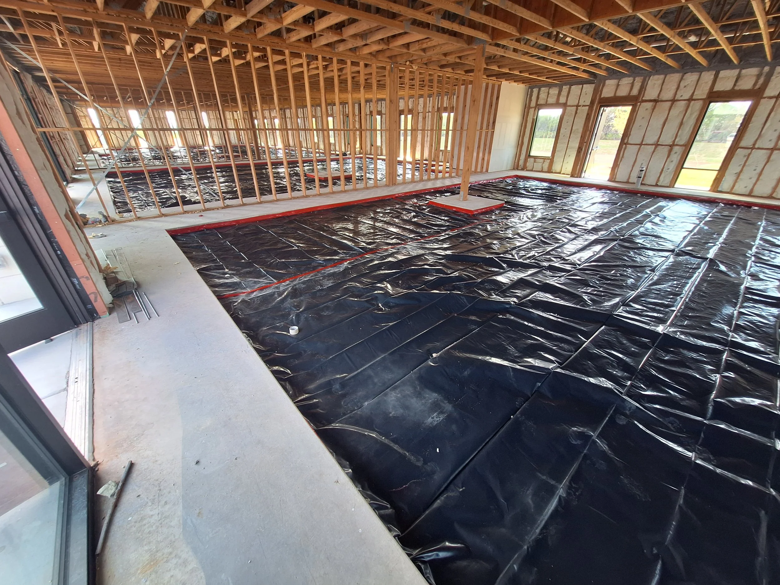Construction site inside a building with exposed wooden framing and black plastic vapor barrier on the floor, with some window and door openings visible.