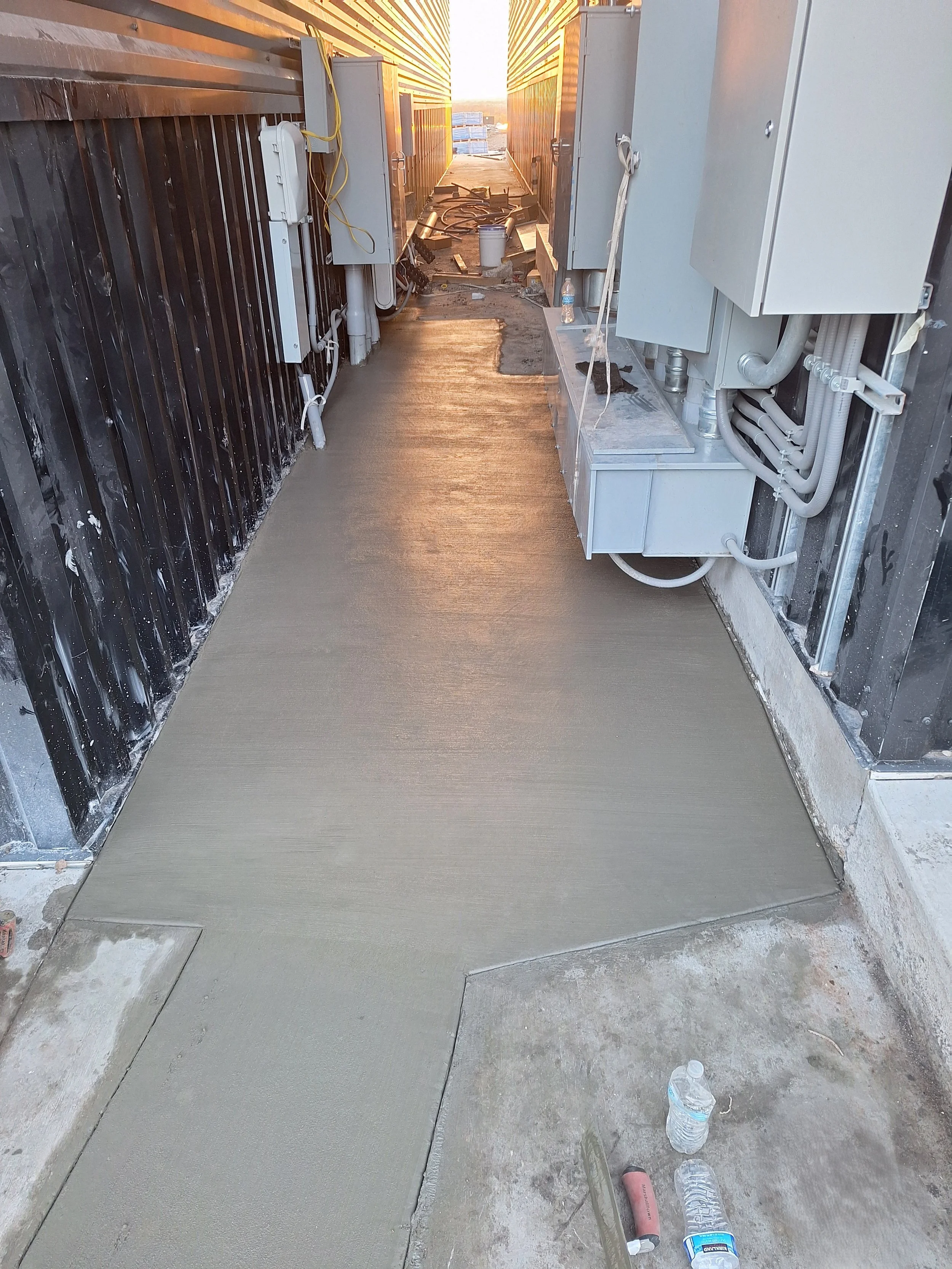 A freshly poured concrete sidewalk between two building structures at sunset. Construction tools and supplies are visible on the ground.