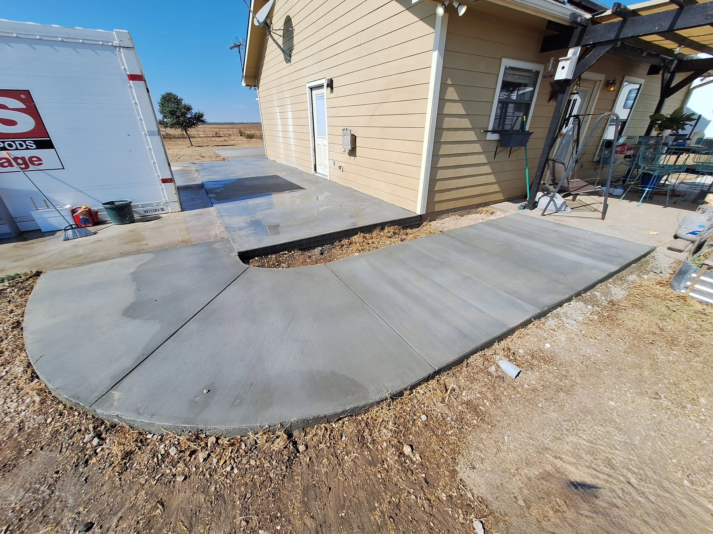 Newly poured concrete sidewalk and ramp leading to the back door of a beige house, with a small patio and outdoor furniture on the right, and a large white storage or moving truck on the left, on a sunny day.
