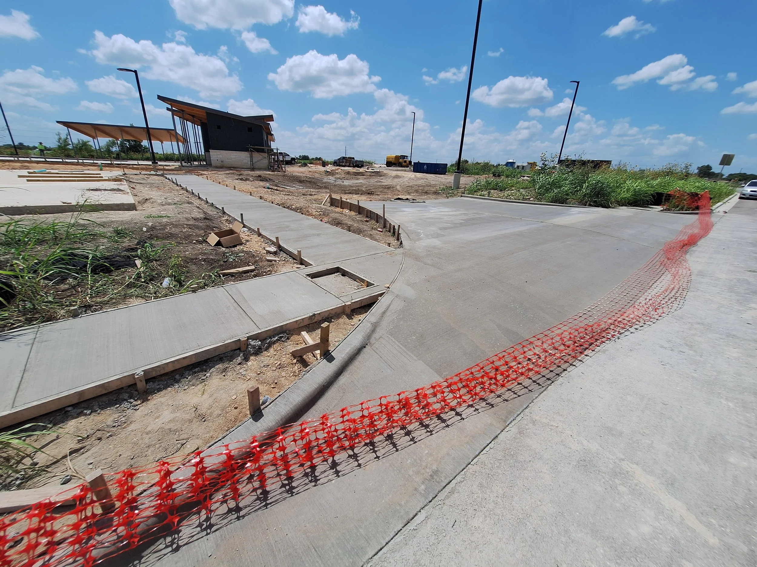 Sidewalk under construction next to an unfinished ramp, with orange safety fencing along the edge, on a sunny day with scattered clouds.
