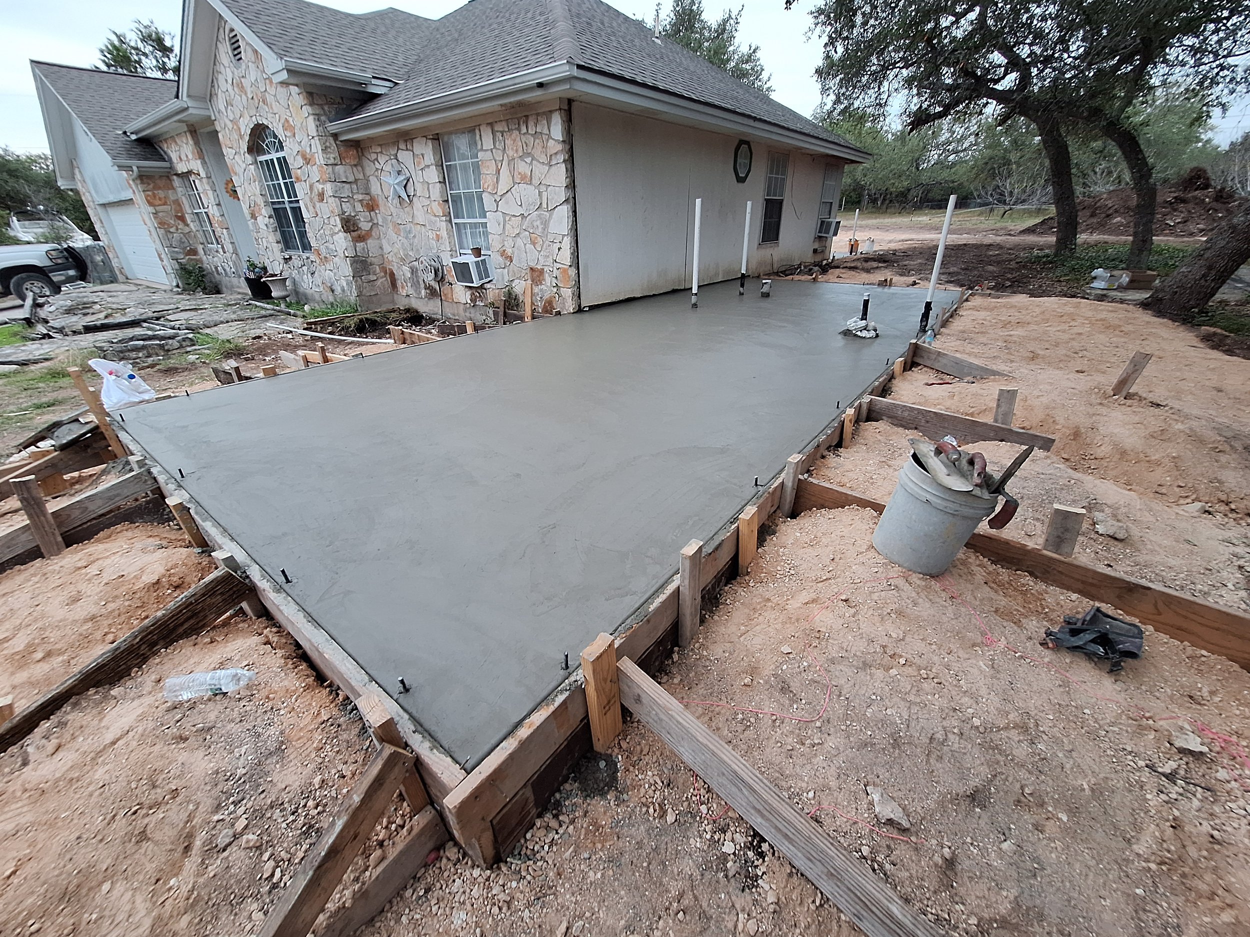 Concrete slab foundation poured in outdoor yard near house with stone and siding exterior, construction tools and materials nearby, and trees in the background.