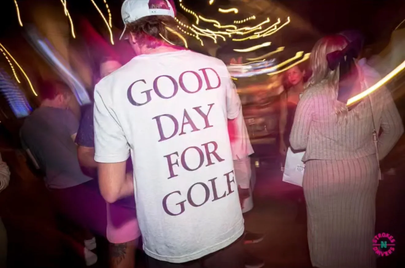 Person wearing a white T-shirt with purple text that reads 'Good Day for Golf,' surrounded by other people in a dimly lit, lively setting with colorful lights.