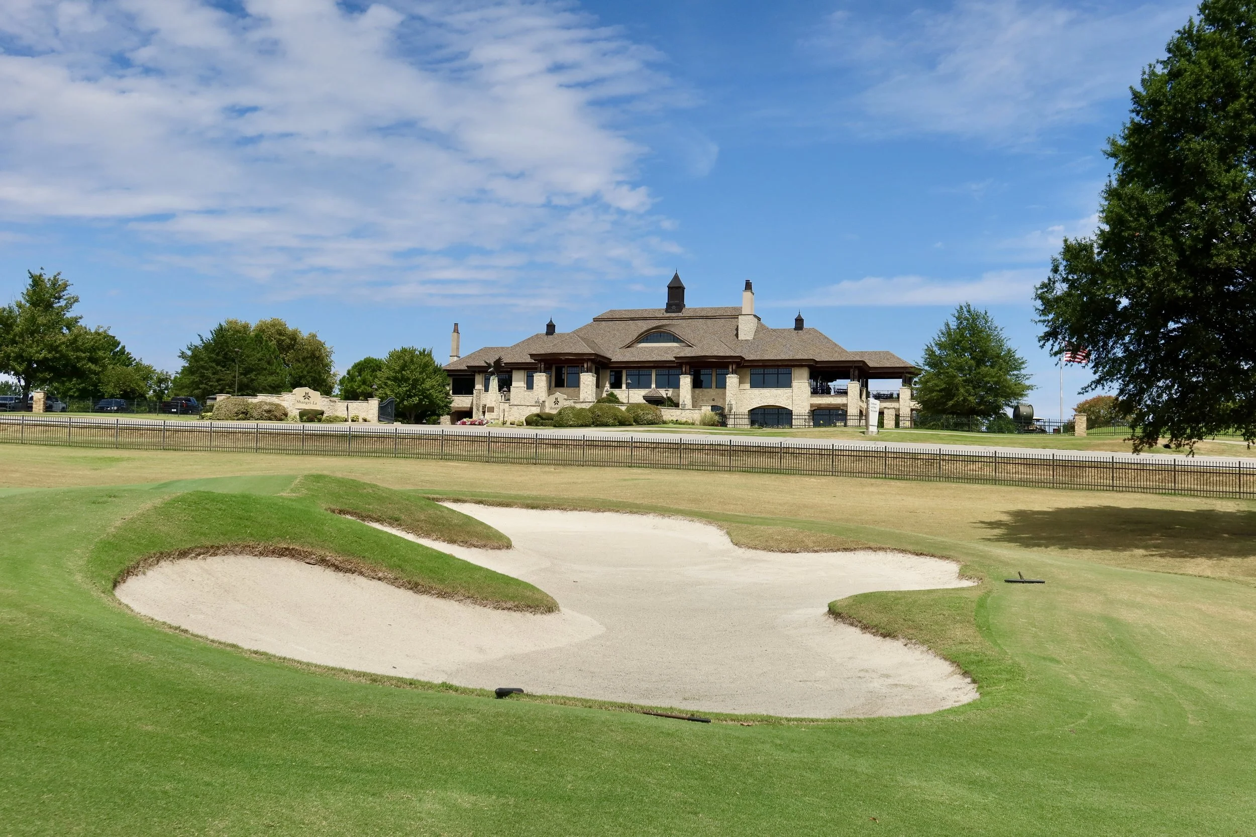 A golf course with a sand trap in the foreground and a large building in the background under a partly cloudy blue sky.