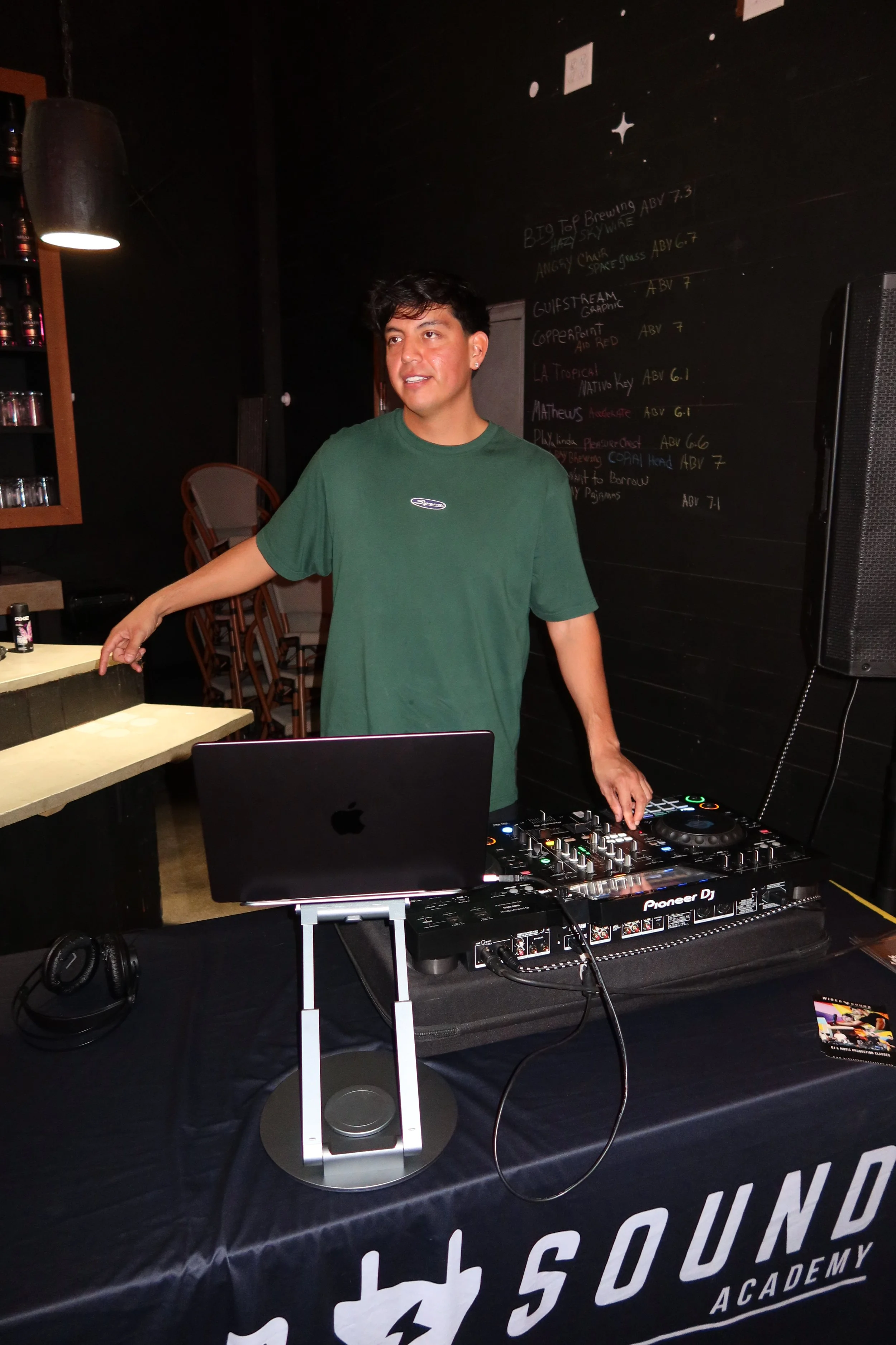 A young man is DJing at a music event, standing behind a DJ controller and a laptop, with a blackboard menu in the background.