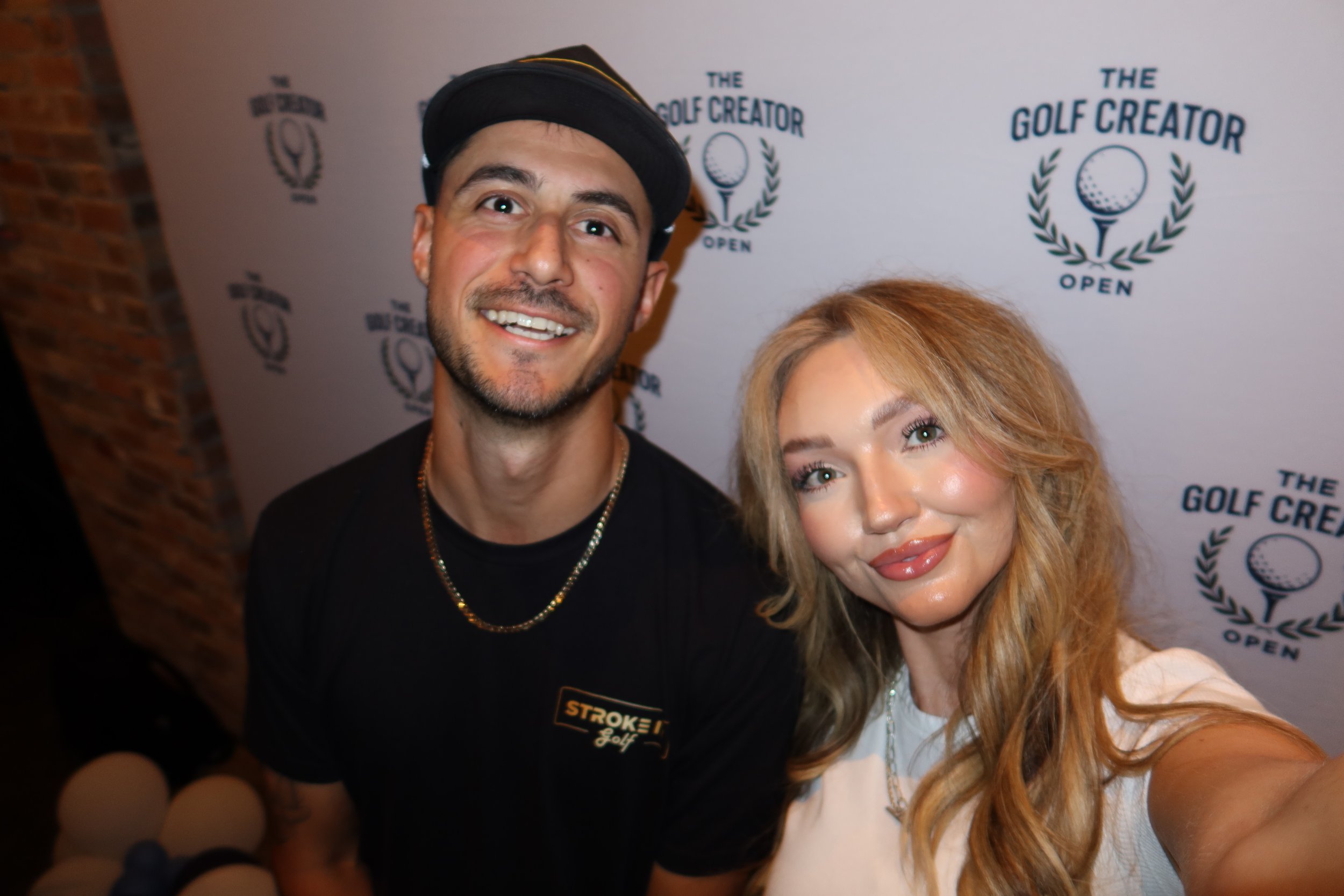 A man and a woman taking a selfie at a golf event, with a backdrop displaying the logo 'The Golf Creator Open' multiple times.