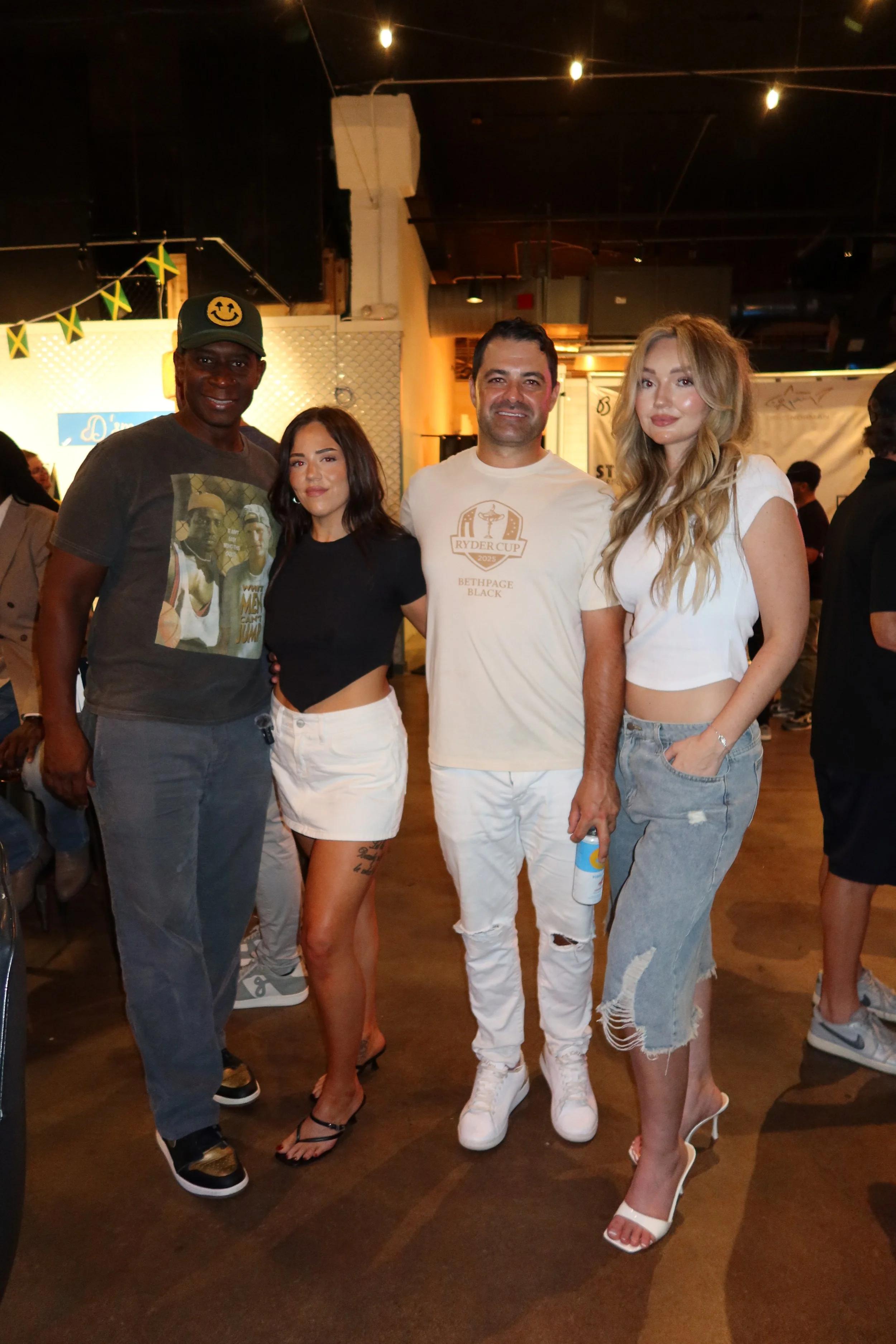 Four people standing together at an indoor event, with a black man wearing a cap with a smiley face, a woman with dark hair in a black crop top and white skirt, a man in a beige t-shirt and white distressed jeans, and a blonde woman in a white crop t