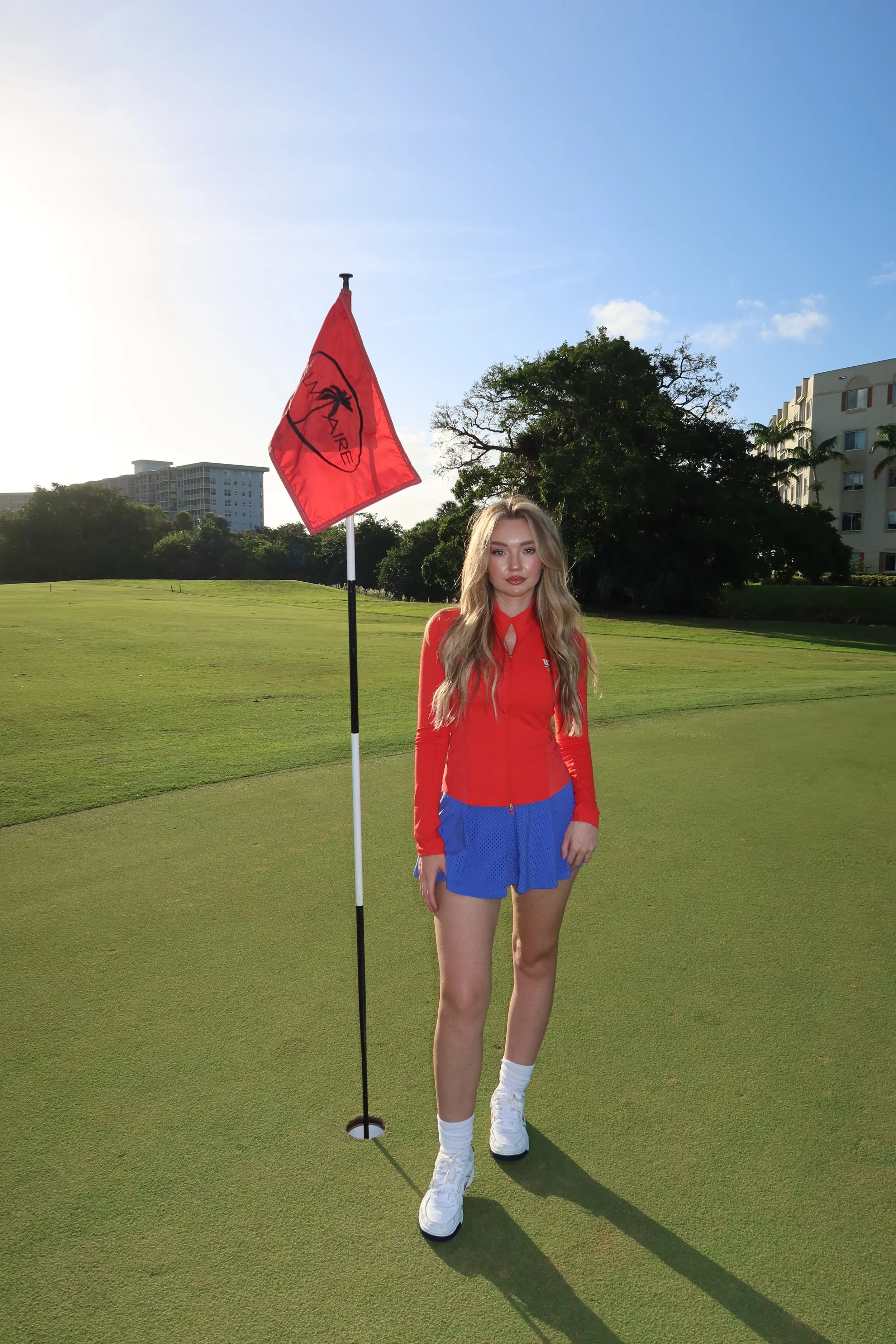 Woman standing on a putting green at a golf course, holding a flagstick with a red flag marked 'LIV'. She is dressed in a red long-sleeve shirt, blue shorts, white socks, and white sneakers, with trees and buildings visible in the background under a 