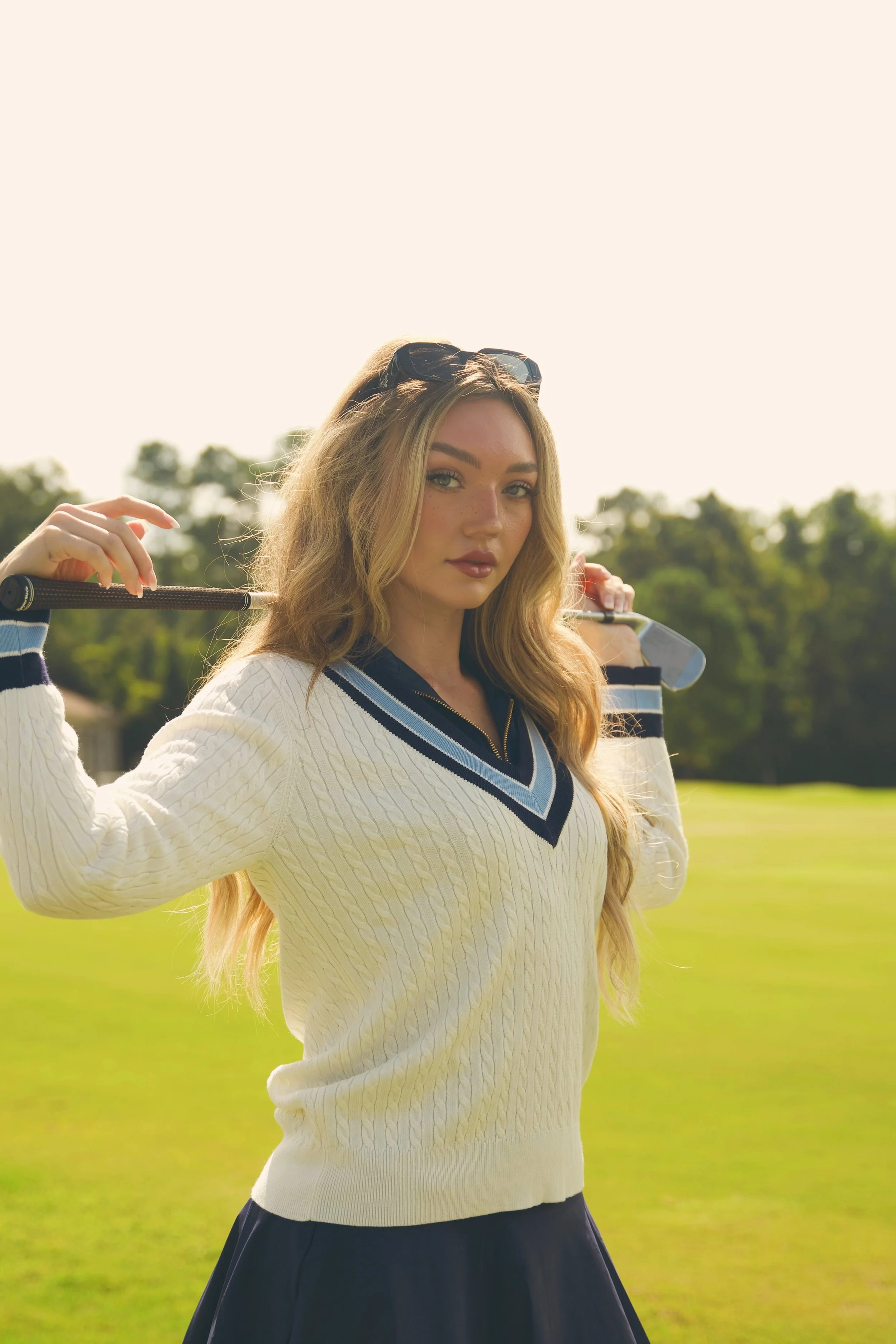 Young woman holding a golf club on a golf course, wearing a white sweater with blue accents and sunglasses on her head.