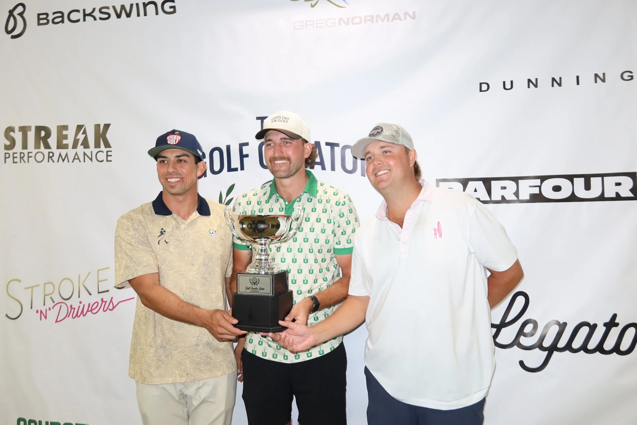 Three men standing together holding a trophy at a golf event, smiling, with a backdrop featuring various sponsor logos including Backswing, Greg Norman, Dunning, and others.