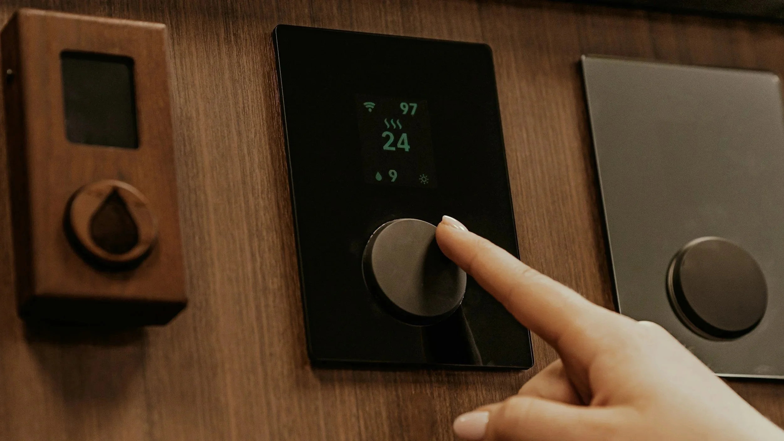 A person's finger adjusting a black digital thermostat on a wooden wall, which displays the temperature, humidity, Wi-Fi signal, and other icons.