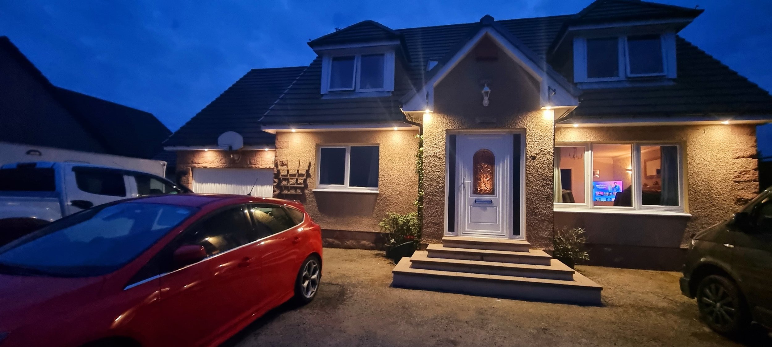A house at night with exterior lights on, three steps leading to the front door, cars parked in the driveway, and a television visible through the living room window.