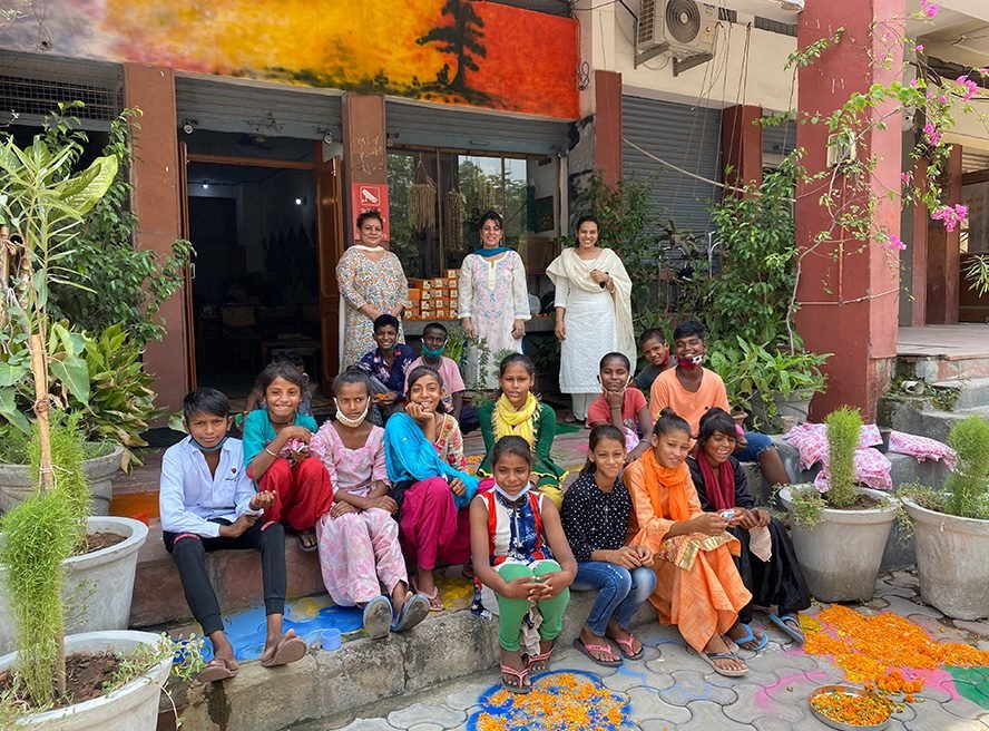 A group of children and three women outside a building, posing for a photo during a celebration with flower decorations on the ground, potted plants, and a colorful mural on the wall.