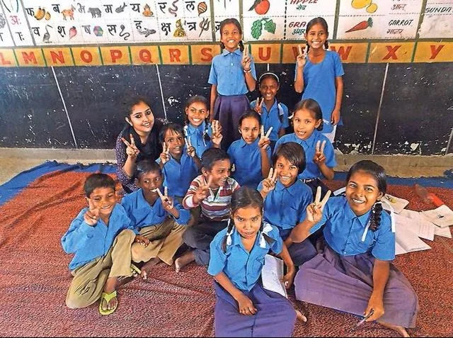 Group of 12 young students in blue uniforms sitting and standing on a classroom rug, making peace signs, with a colorful alphabet chart and drawings on the wall behind them.