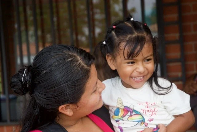 A woman holding a young girl with pigtails, both smiling, outside near a fence and brick wall.