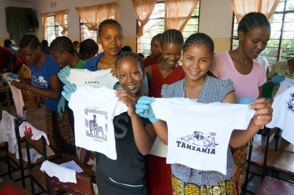 Group of young Tanzanian girls in a classroom holding up white T-shirts with Africa and Tanzania printed on them, all wearing gloves and smiling.