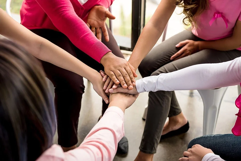 Multiple people with hands stacked together in a circle, symbolizing unity and support, in a bright indoor setting.
