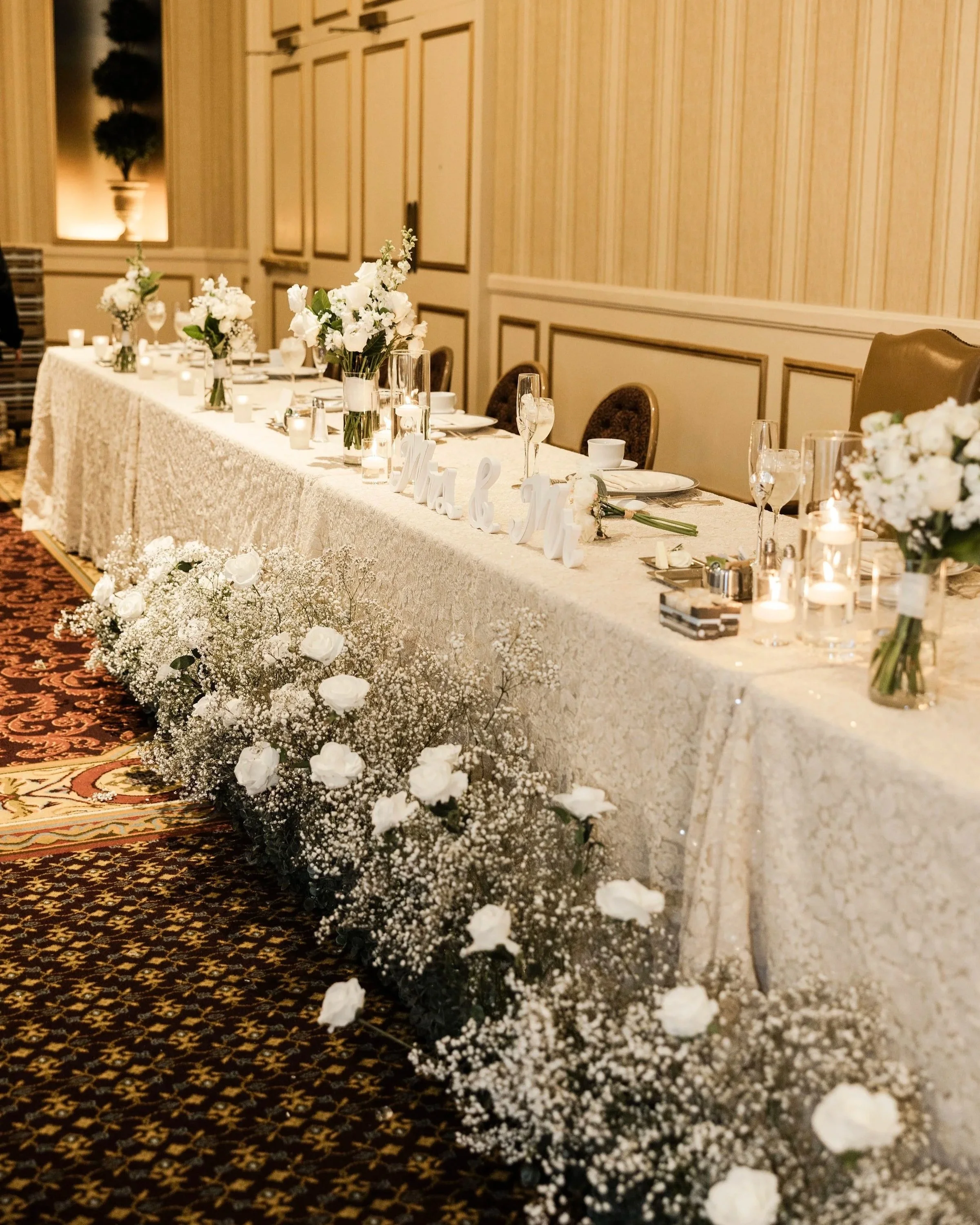 Wedding reception table decorated with white flowers, candles, and elegant table settings in a banquet hall.