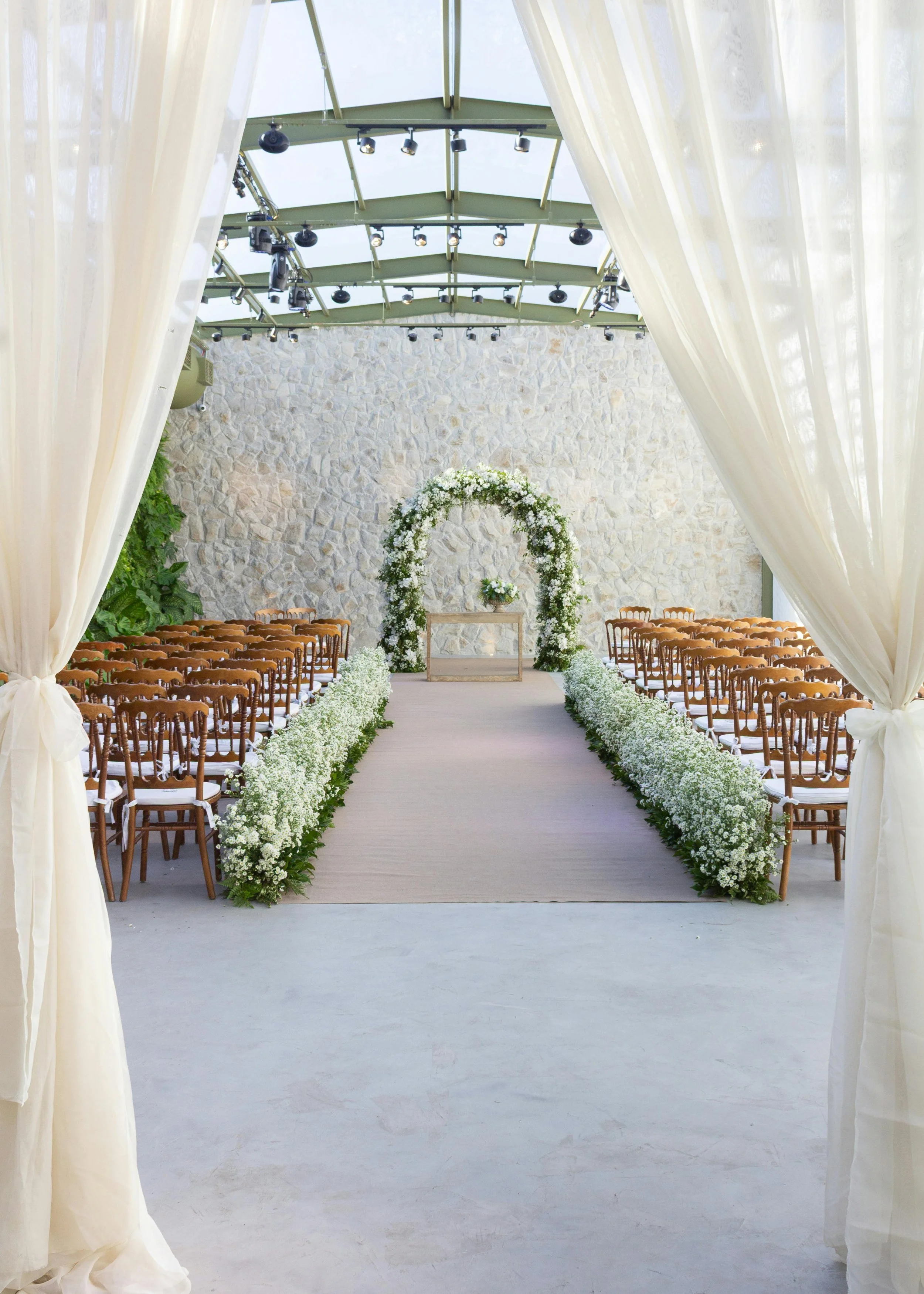 Indoor wedding ceremony setup with chairs on either side of an aisle, floral arch at the end, and sheer curtains framing the entrance.