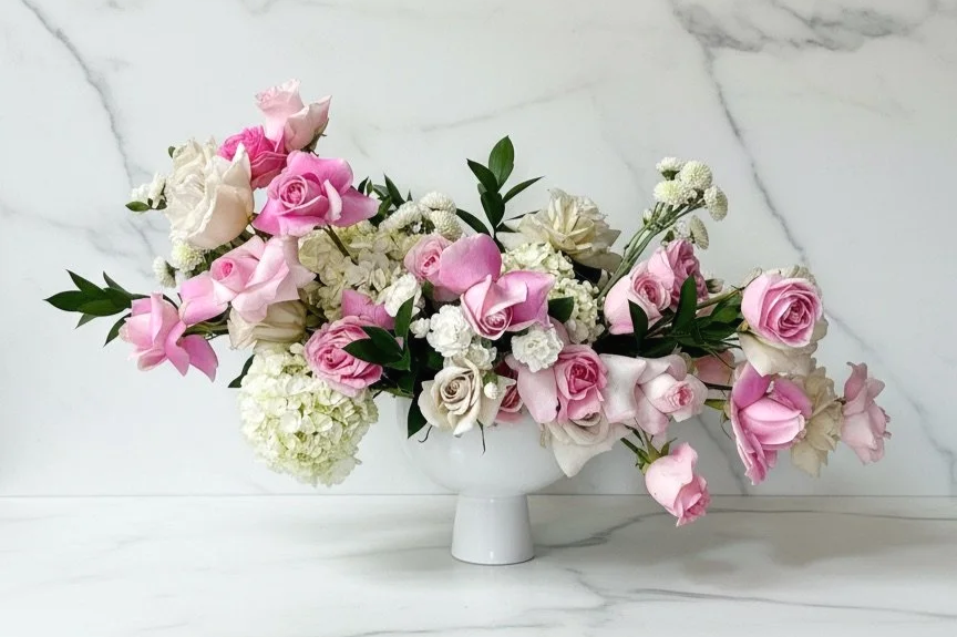A white vase filled with pink and white roses, white hydrangeas, and small white filler flowers on a marble surface.