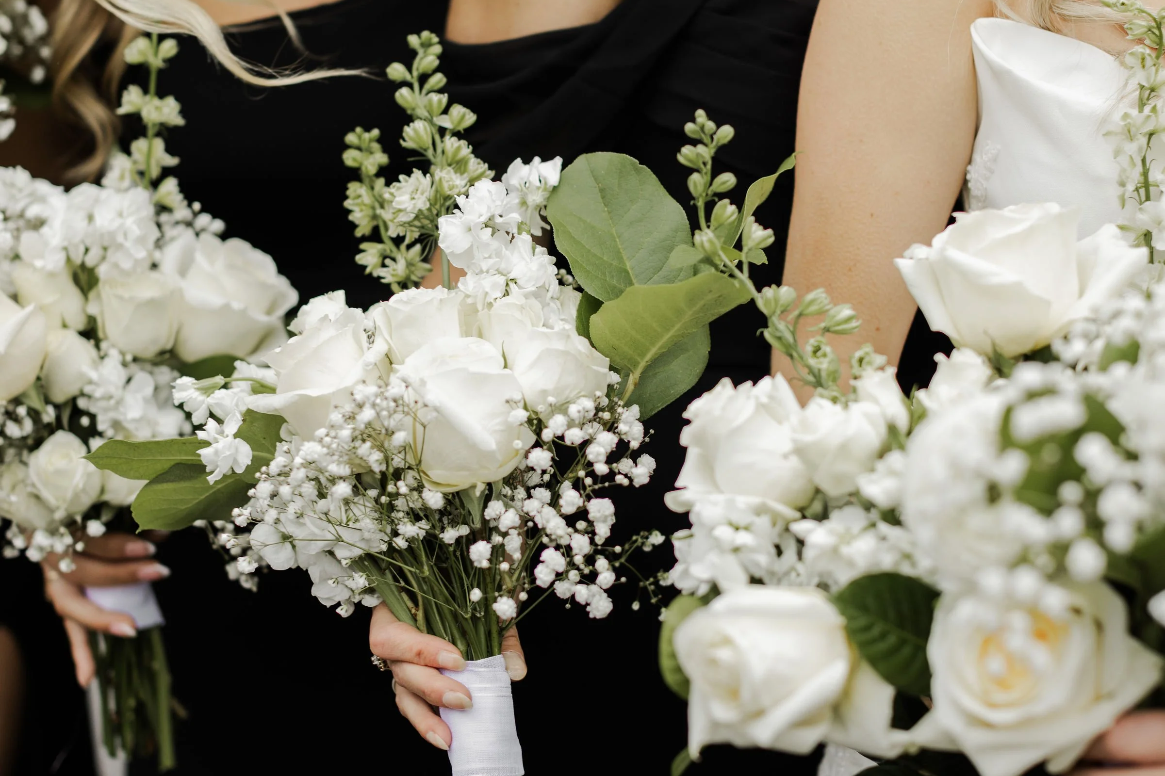 Close-up view of white bouquets of roses, baby's breath, and greenery, held by women dressed in black and white dresses, likely at a wedding.