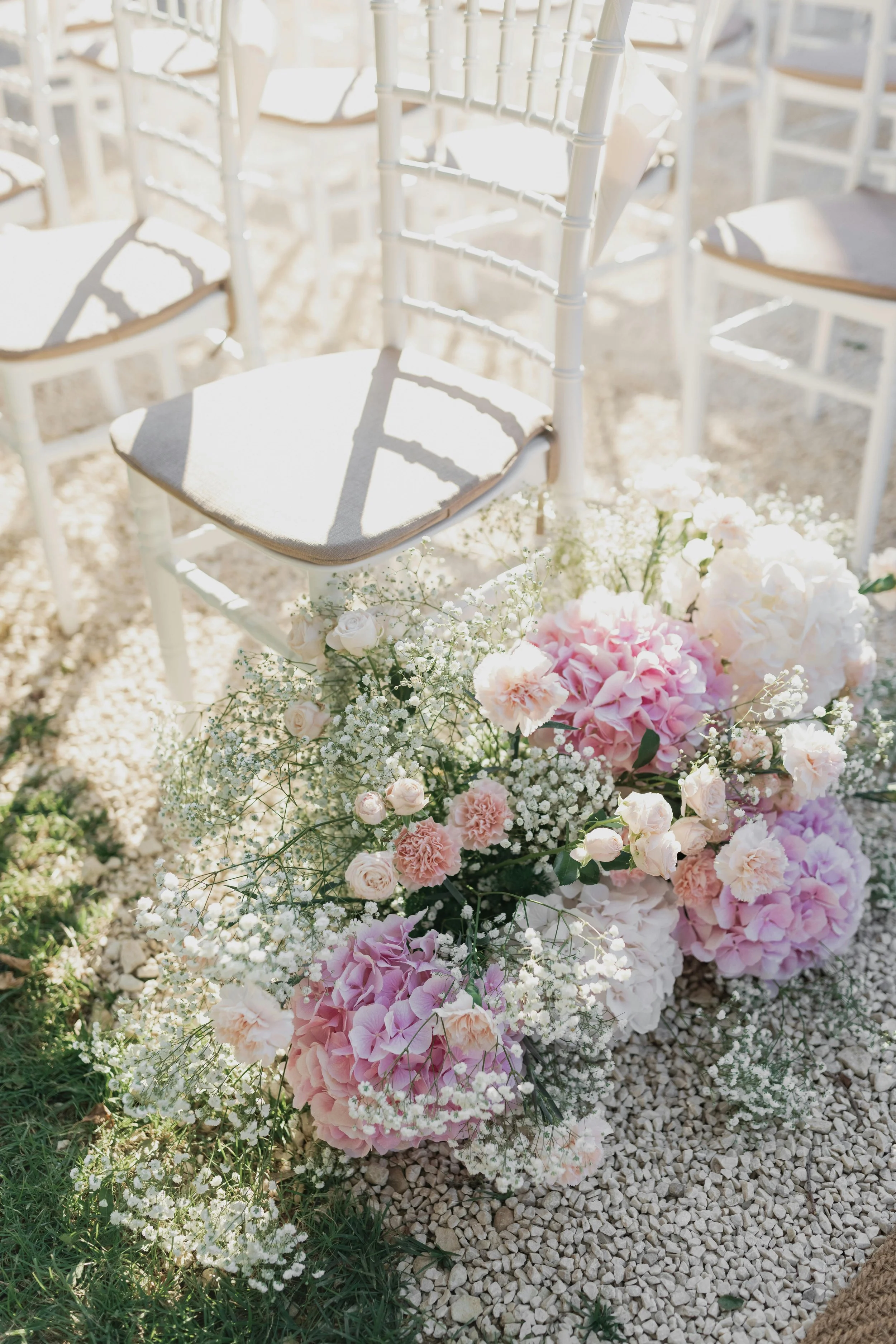 White chairs with beige cushions arranged outdoors on gravel, decorated with pink and white flowers, at a wedding or outdoor event.