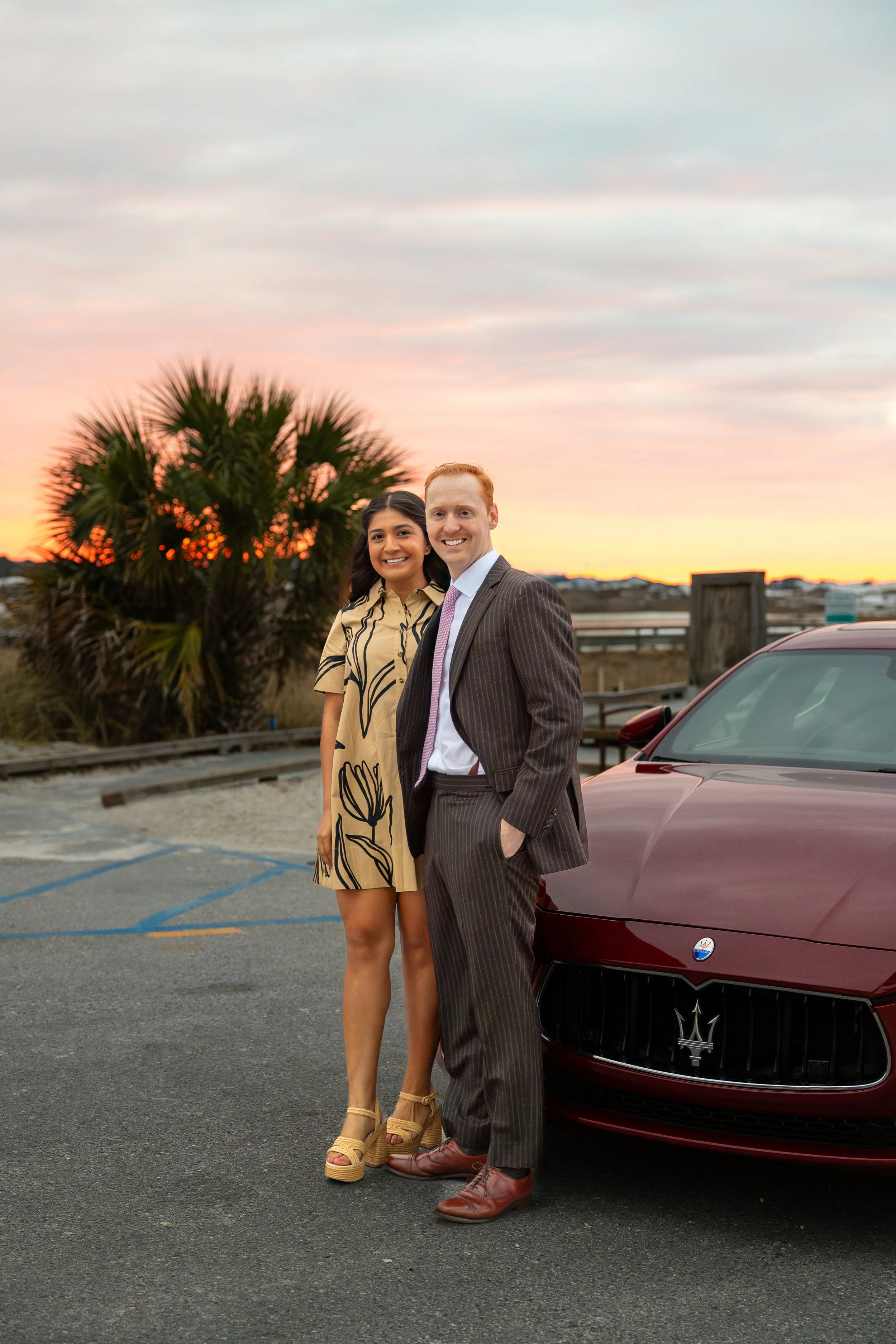 A man and a woman standing next to a red Maserati car in an outdoor parking lot at sunset. The woman is wearing a beige dress with black floral patterns and heels, while the man is dressed in a pinstripe suit with a pink tie. They are smiling and standing close together.