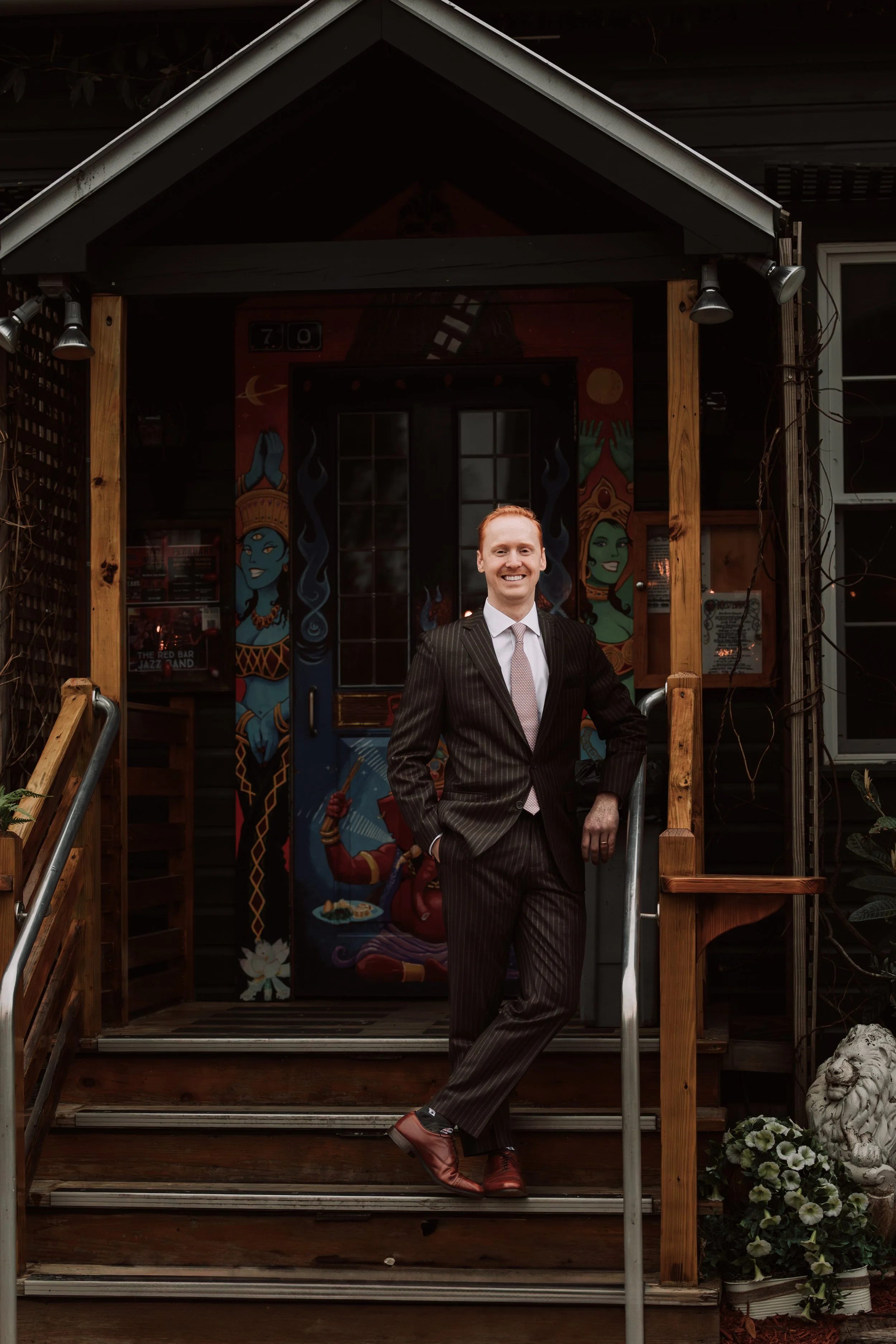 A man in a dark striped suit and tie standing on wooden steps outside a building with painted door and wall art, smiling at the camera.