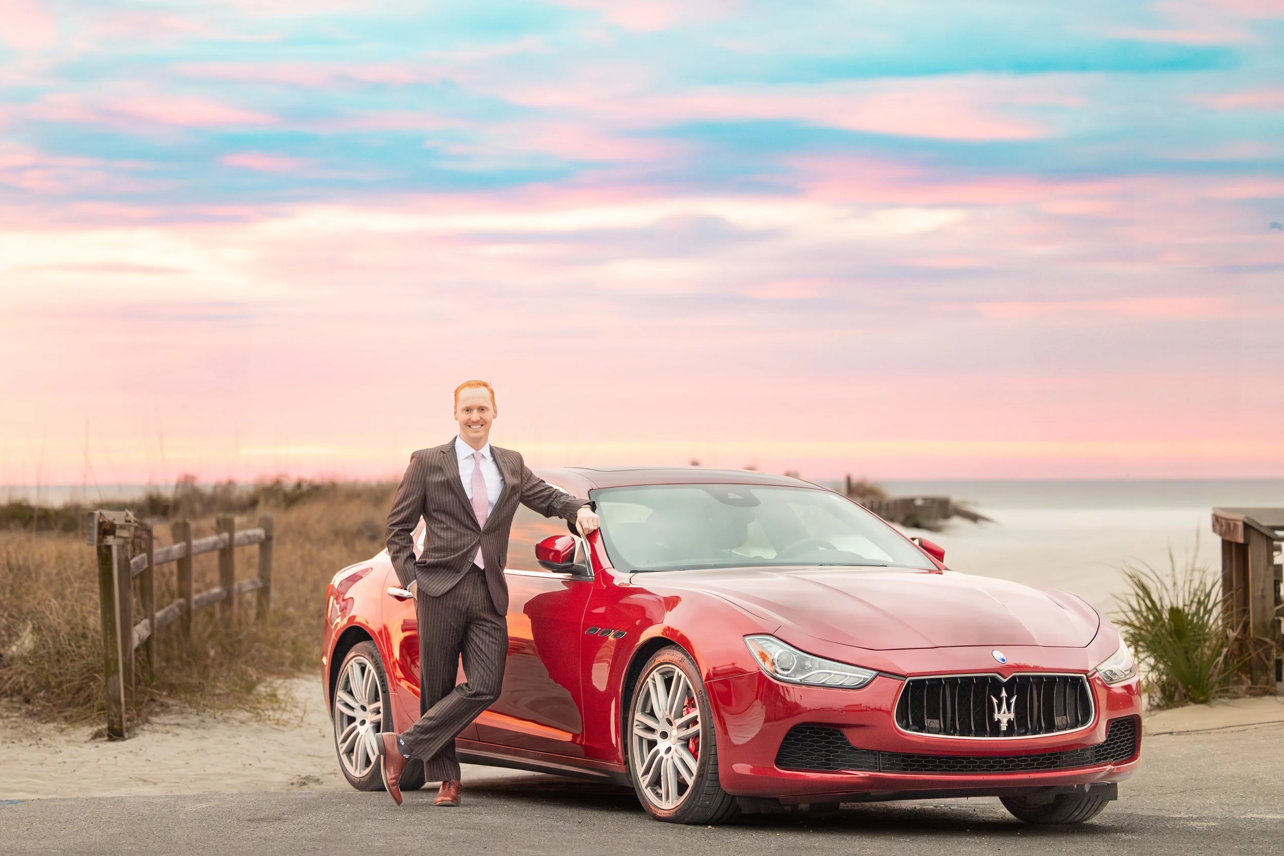 A man in a suit leaning on a red Maserati convertible car on a beach road during sunset.