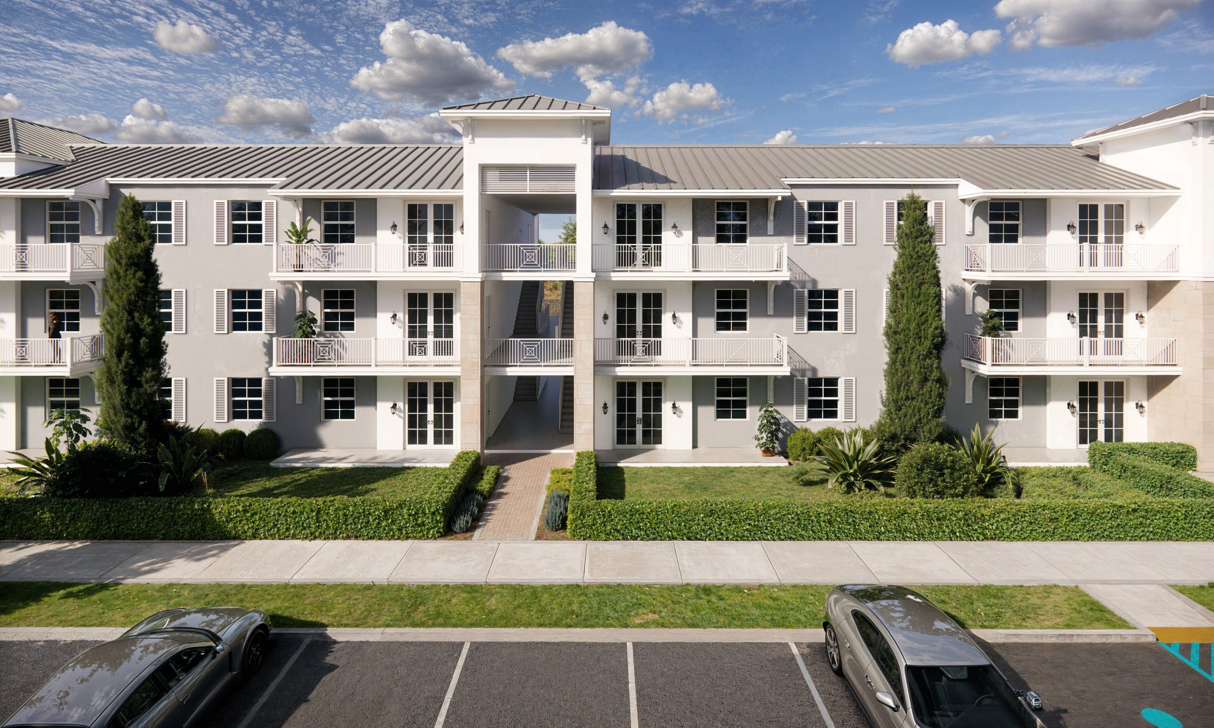 Front view of a modern three-story apartment building with white exterior walls, multiple balconies with white railings, tall green trees, landscaping, and parked cars in the parking lot.