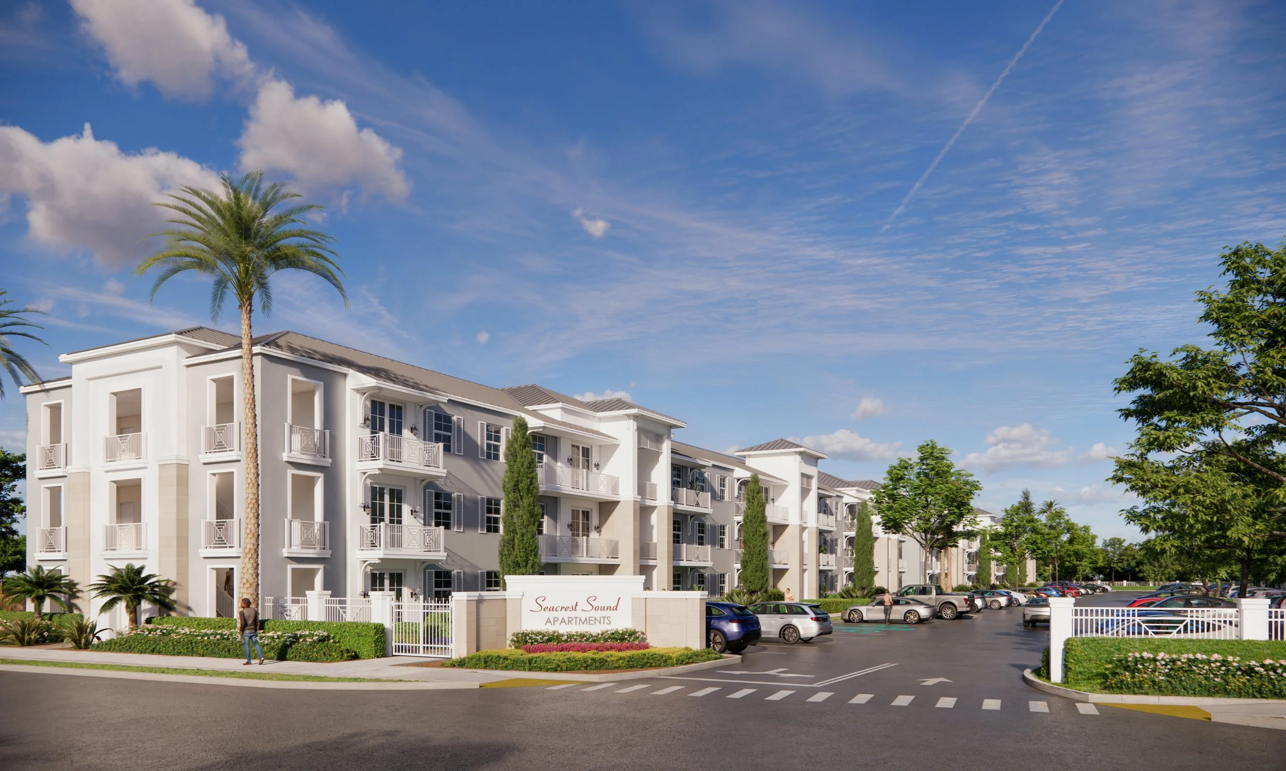 Exterior view of Seacrest Sound Apartments with a white building, palm trees, green shrubbery, parking lot, and blue sky with clouds.