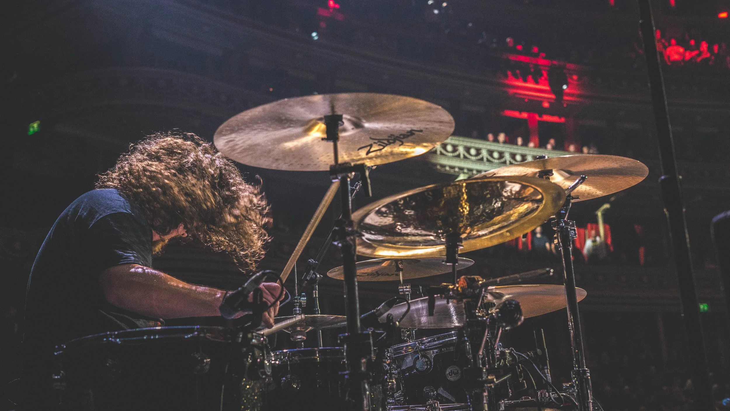 A person with curly hair playing drums on stage with cymbals visible, in a dimly lit venue with red lighting and ornate balcony railing in background.