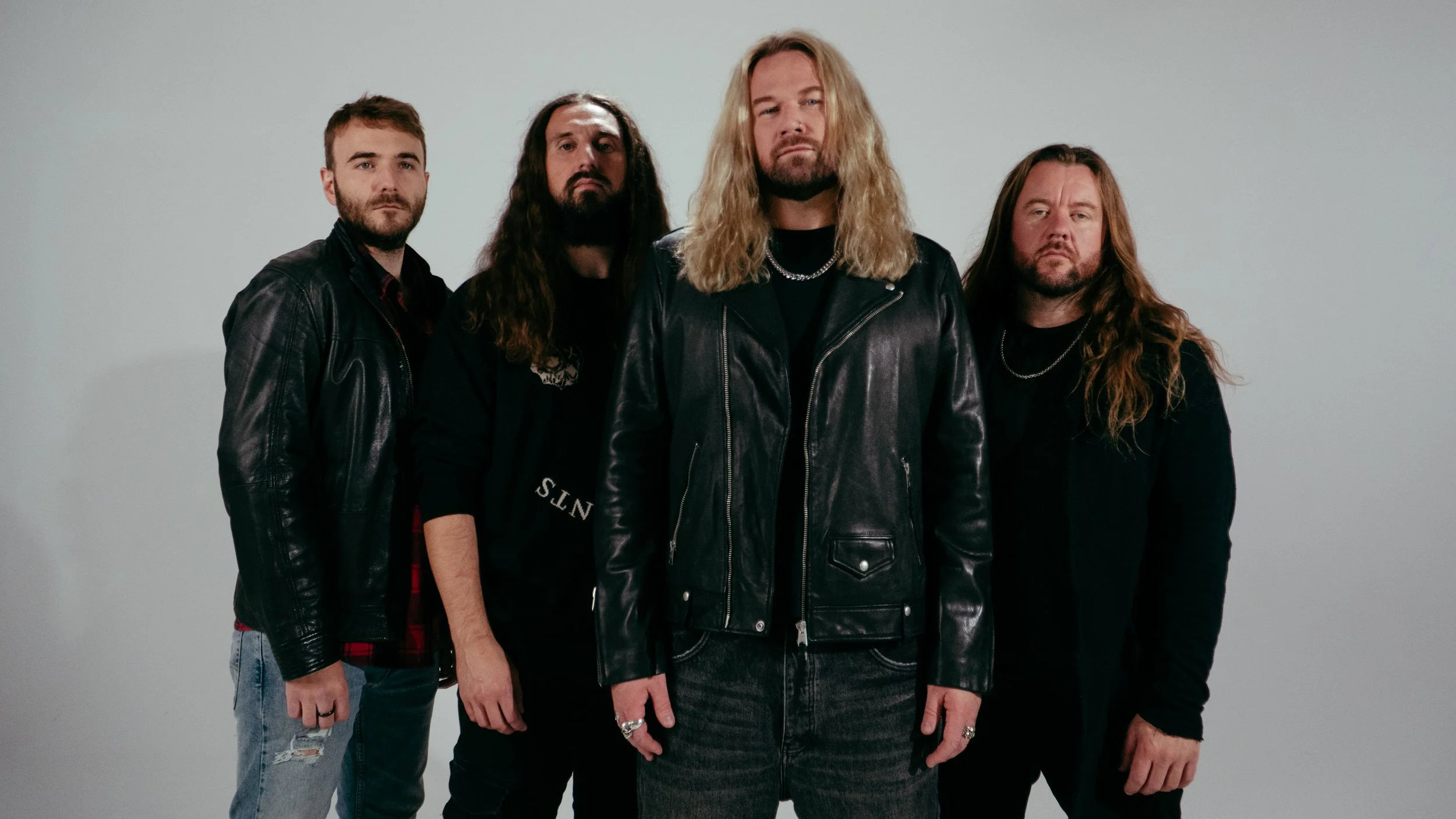 A group of four men standing against a plain white background, wearing dark clothing and leather jackets, with long hair and serious expressions.