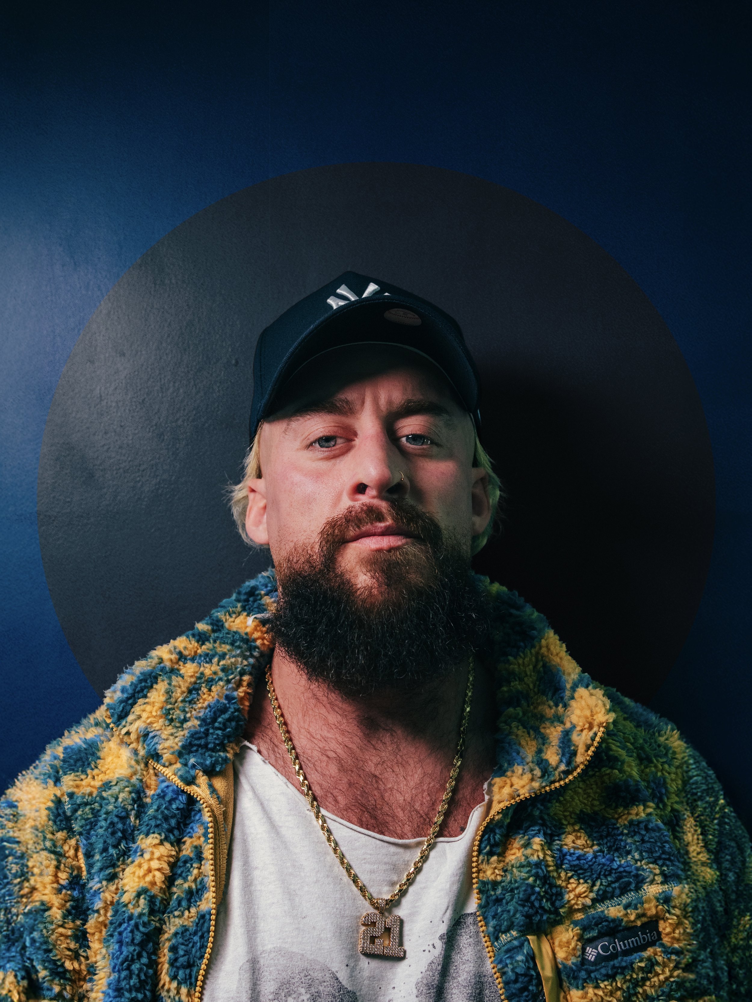 A man with a beard and mustache wearing a black baseball cap, a colorful fleece jacket, a white T-shirt, and gold chains with a pendant that has the number 21. He is standing against a dark wall with a circular design behind his head.