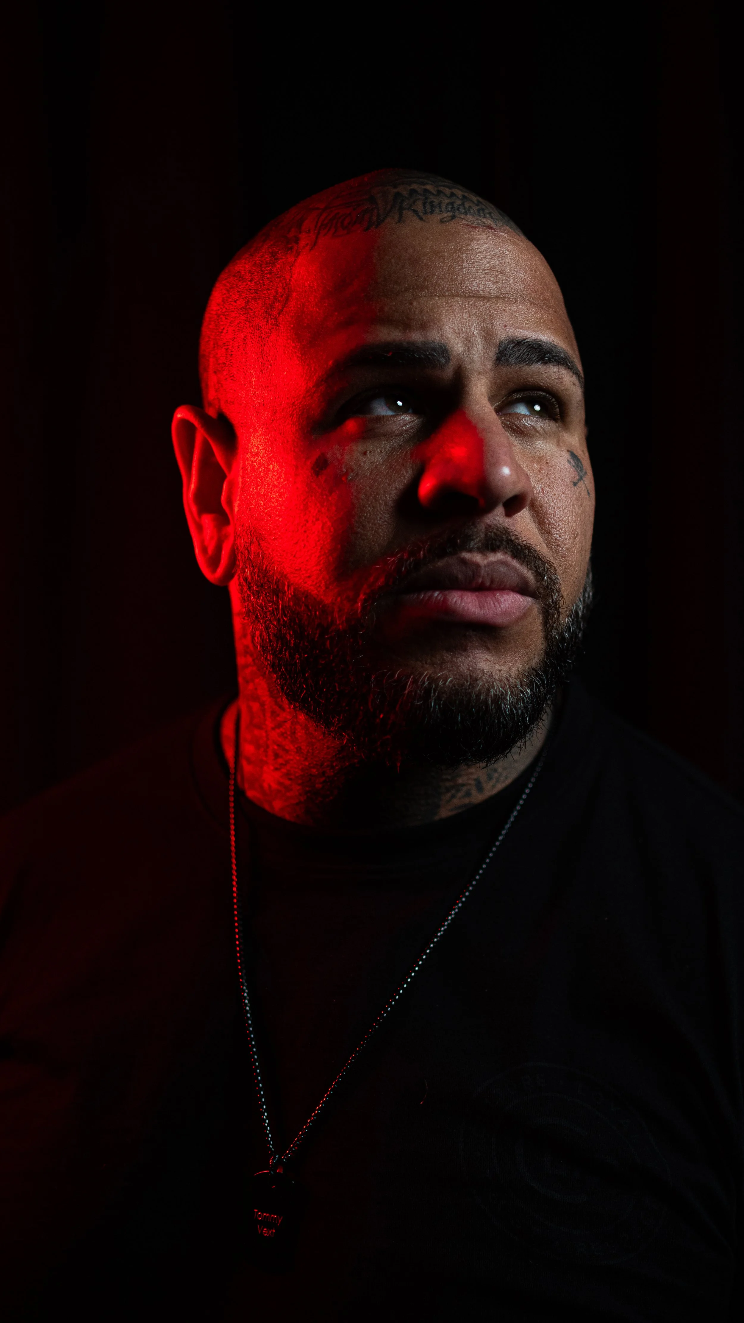 A close-up portrait of a man with tattoos on his head and face, looking upwards with a serious expression, illuminated by red lighting on one side and dark background.