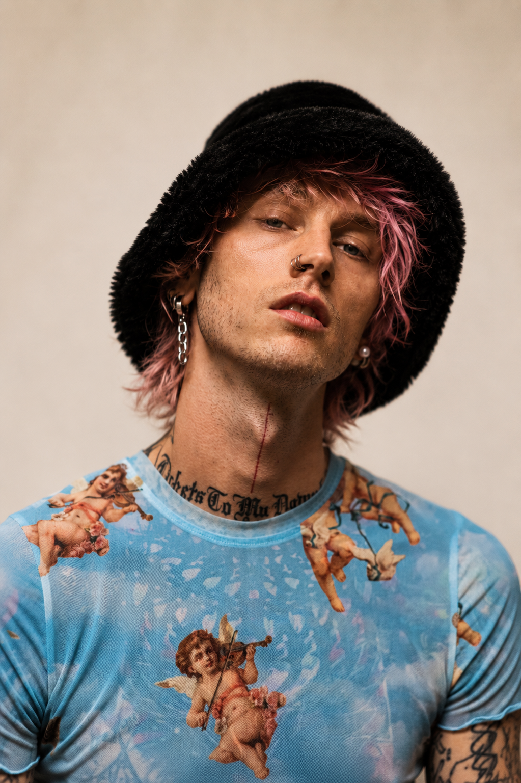 A person with pink hair, wearing a black fuzzy hat, a blue shirt with cherub and angel images, multiple earrings, and tattoos, poses with a serious expression.