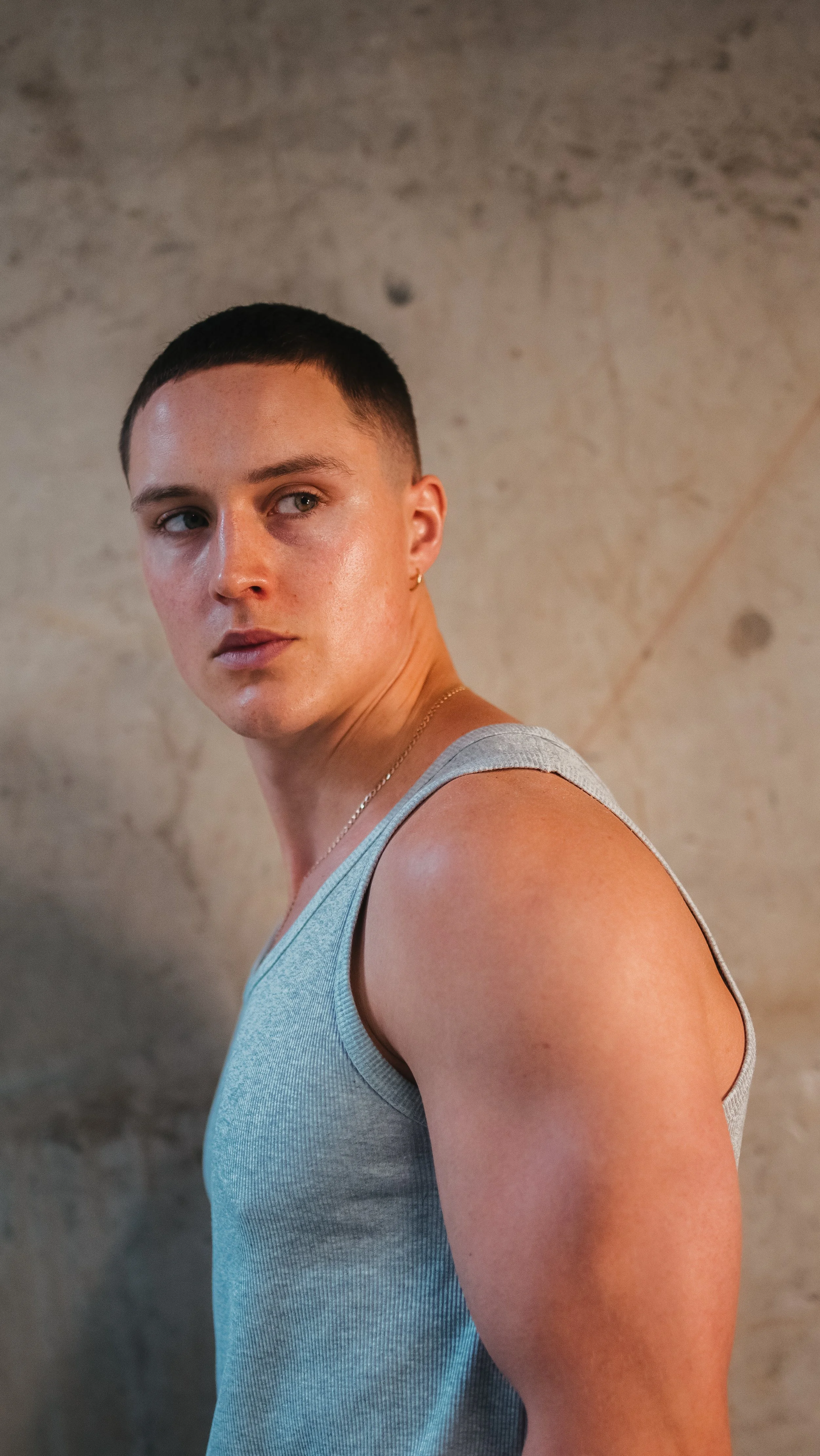 A young man with a short haircut and light skin wearing a light gray sleeveless shirt, standing against a plain beige wall, looking over his shoulder.
