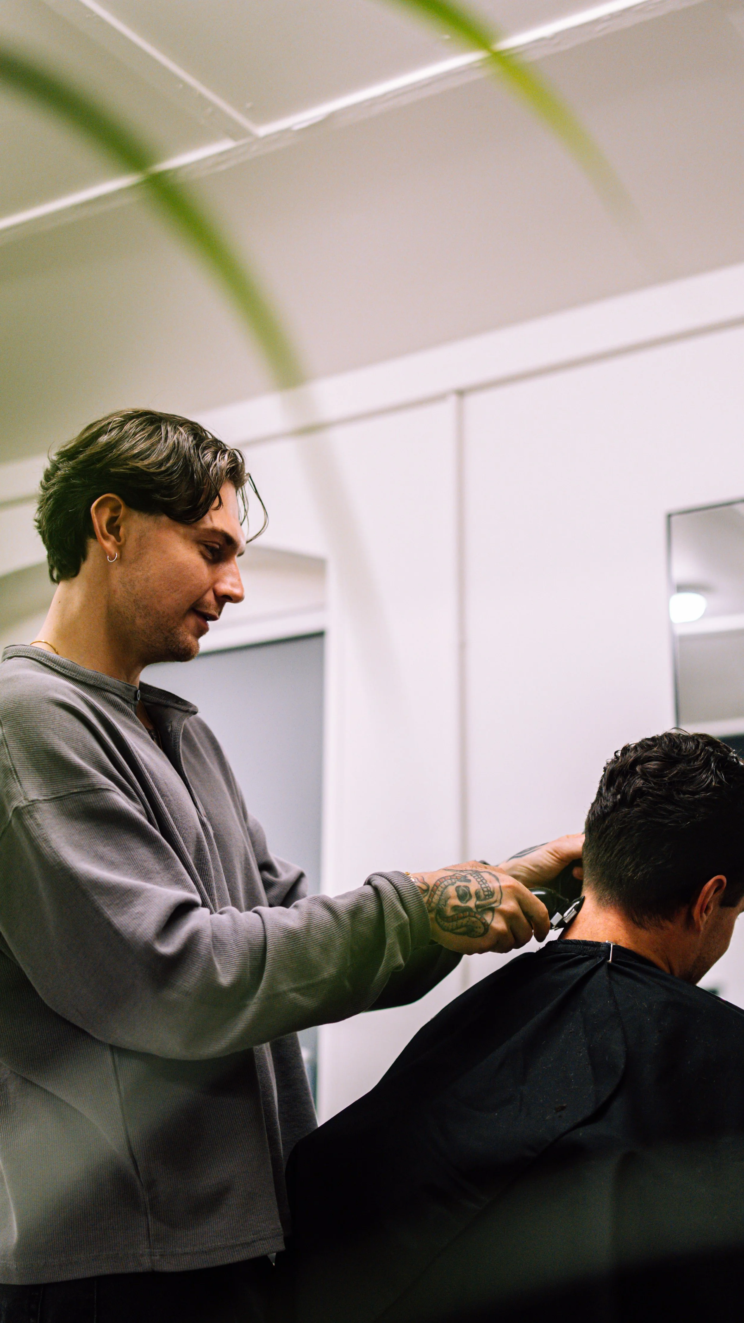 A man with dark hair and tattoos giving a haircut to another man in a salon.