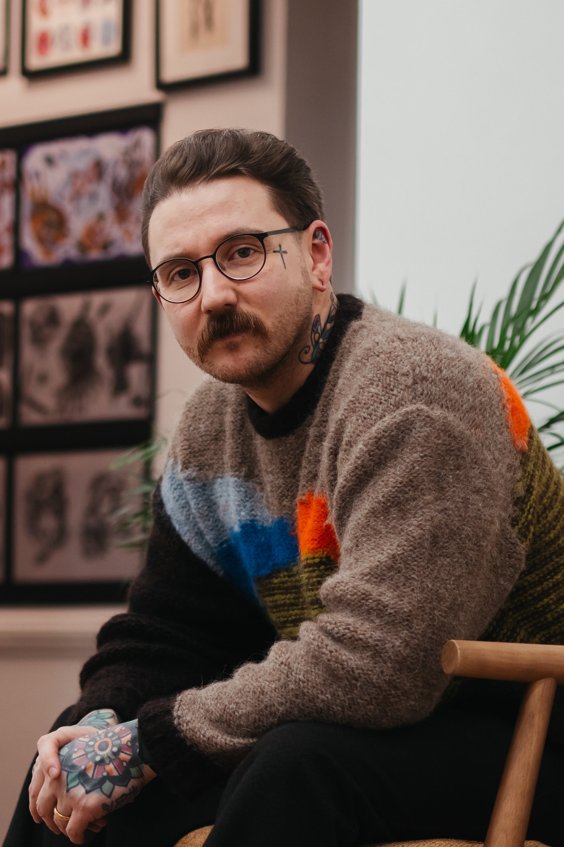 A man with glasses, tattoos, and a mustache wearing a colorful sweater, sitting on a wooden chair, with a wall of framed artwork behind him and a plant to his side.