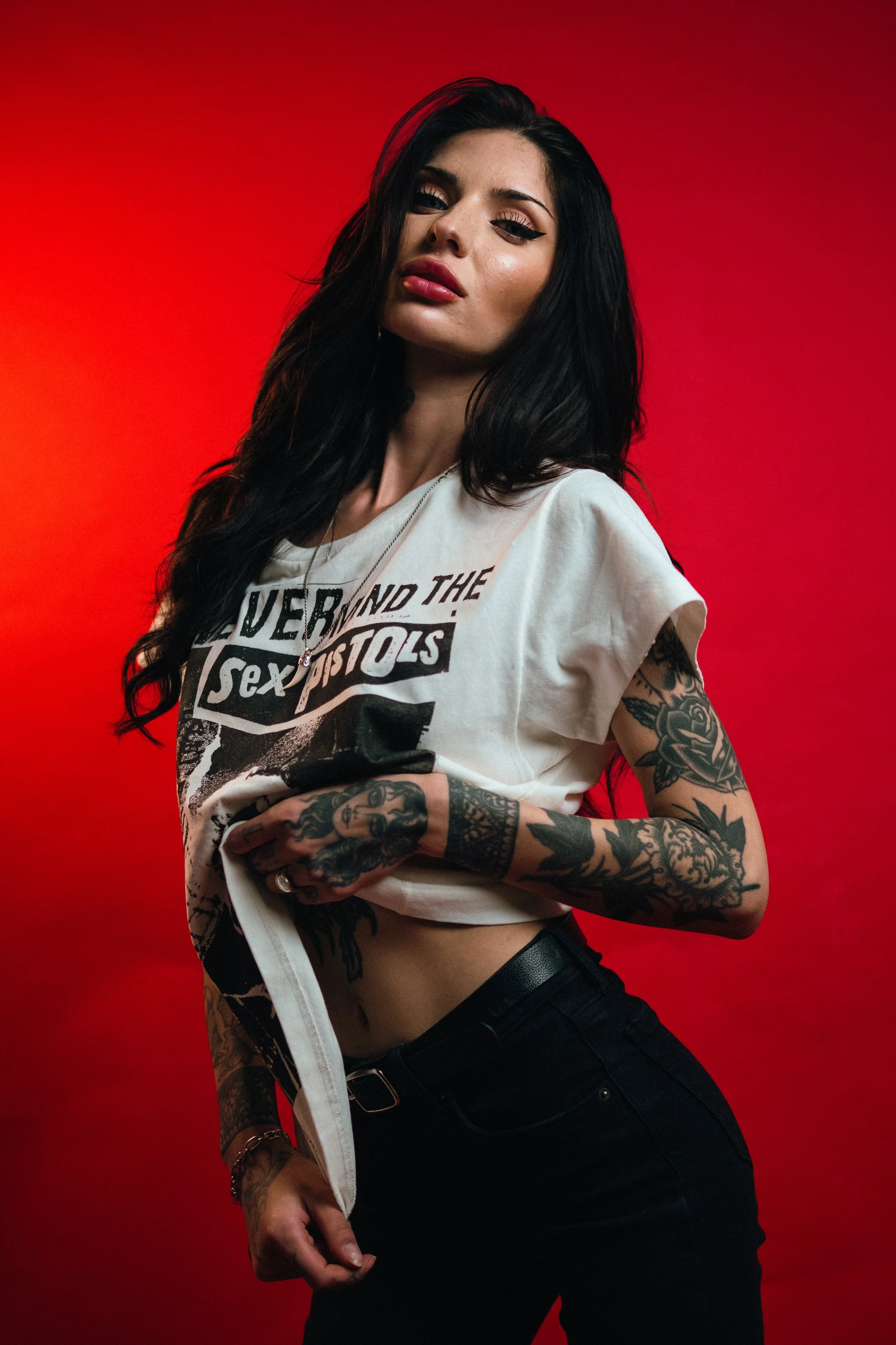 A woman with long black hair and tattoos, wearing a white T-shirt and black pants, stands against a red background.