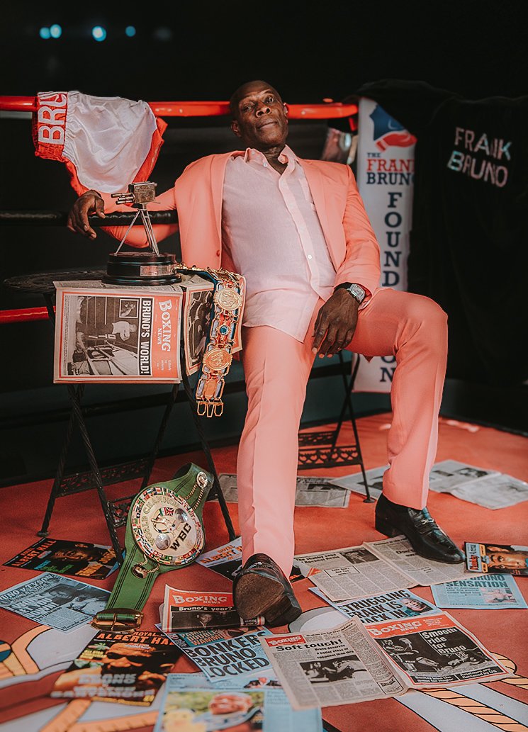 A man in a salmon-colored suit sitting in a boxing ring, surrounded by boxing memorabilia, newspapers, and awards, with a trophy and championship belts on the floor.