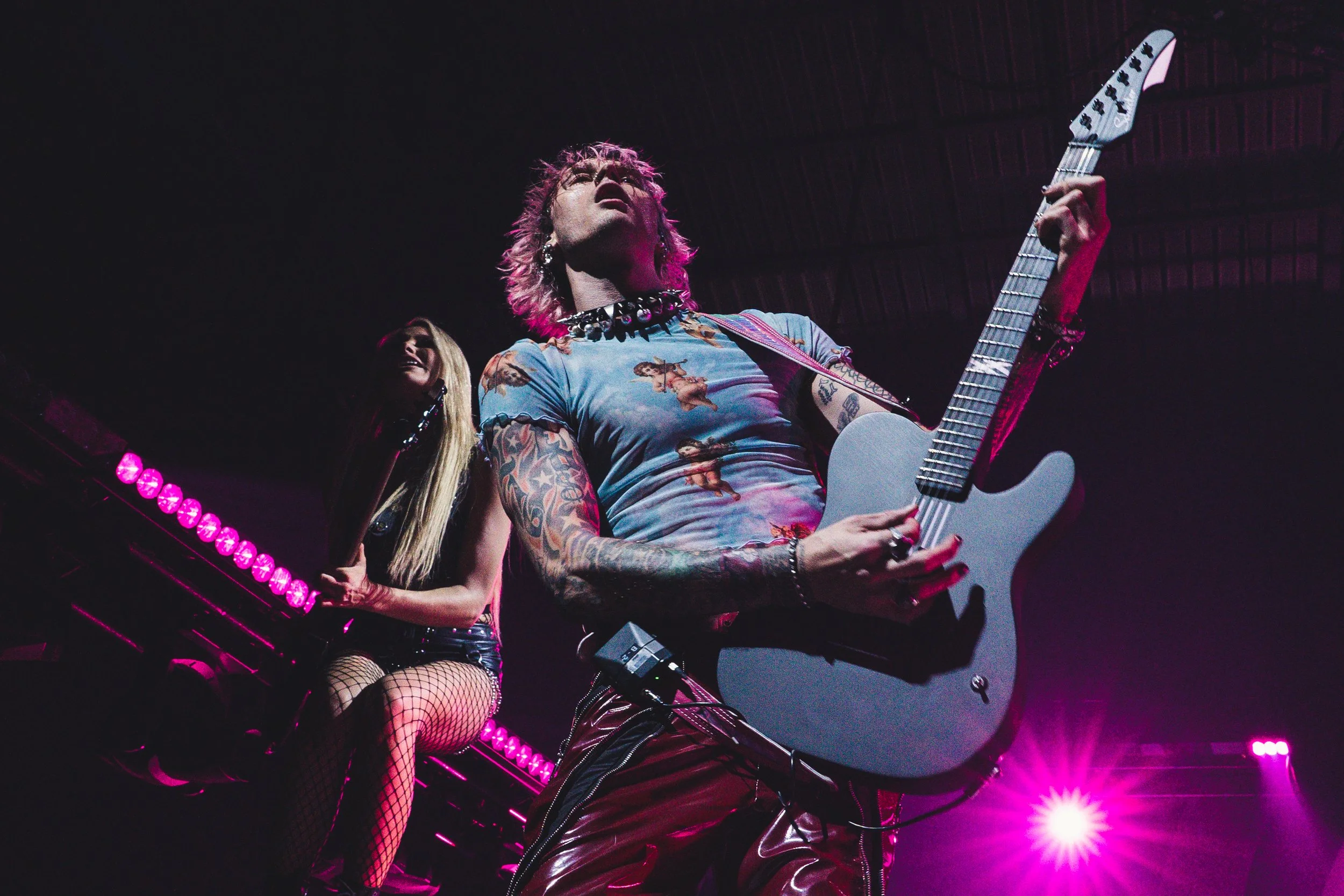 Musicians performing on stage with pink and purple lighting, one playing an electric guitar and the other sitting with a drumstick.