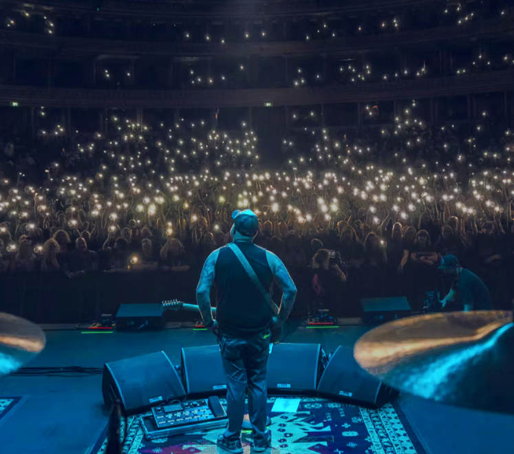 Musician on stage performing at a concert with an audience holding up smartphones with flashlight. Large venue with multiple floors.