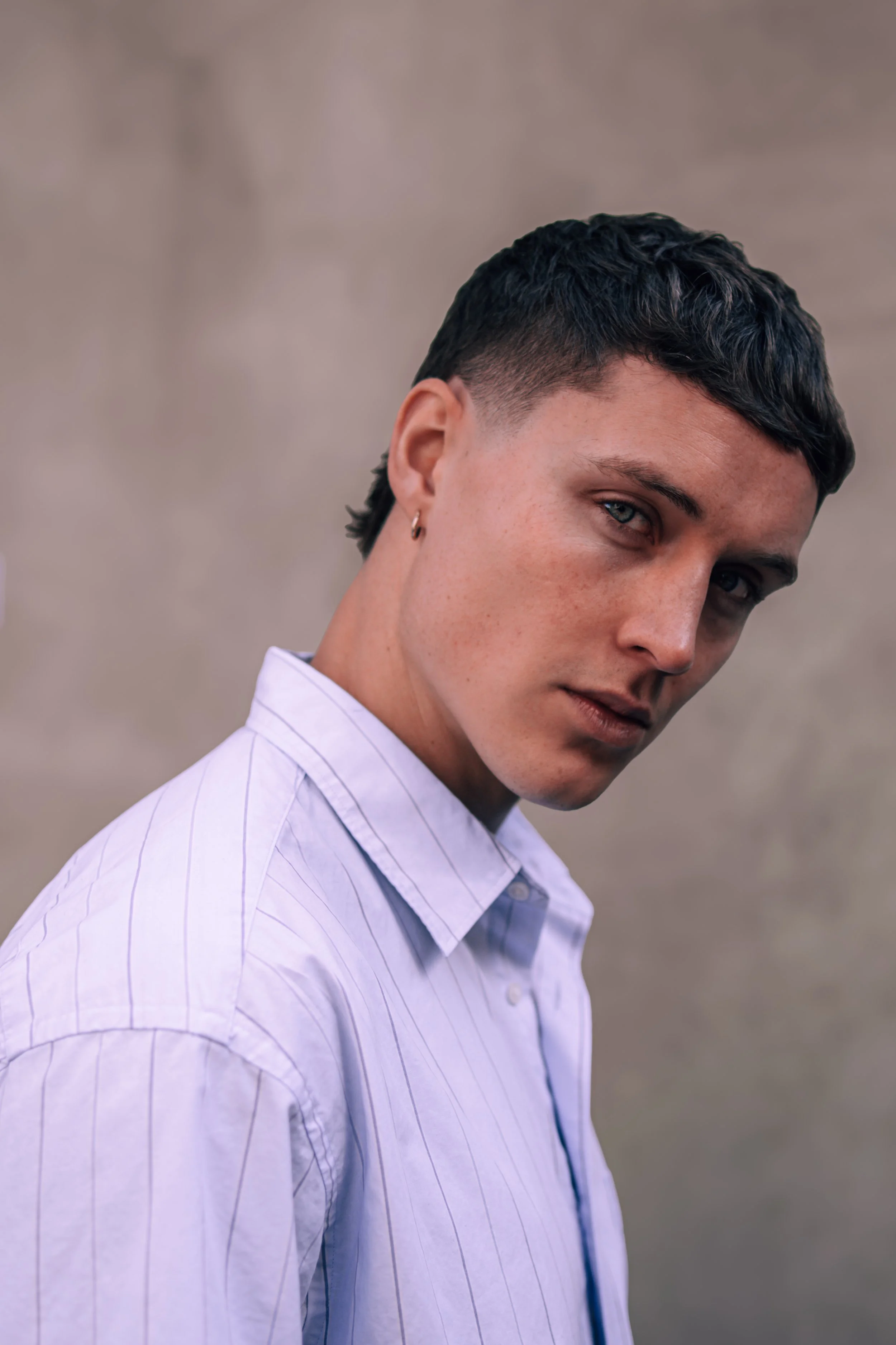 Portrait of a young person with short dark hair, wearing a light blue striped shirt, looking slightly to the side with a neutral expression, against a neutral background.