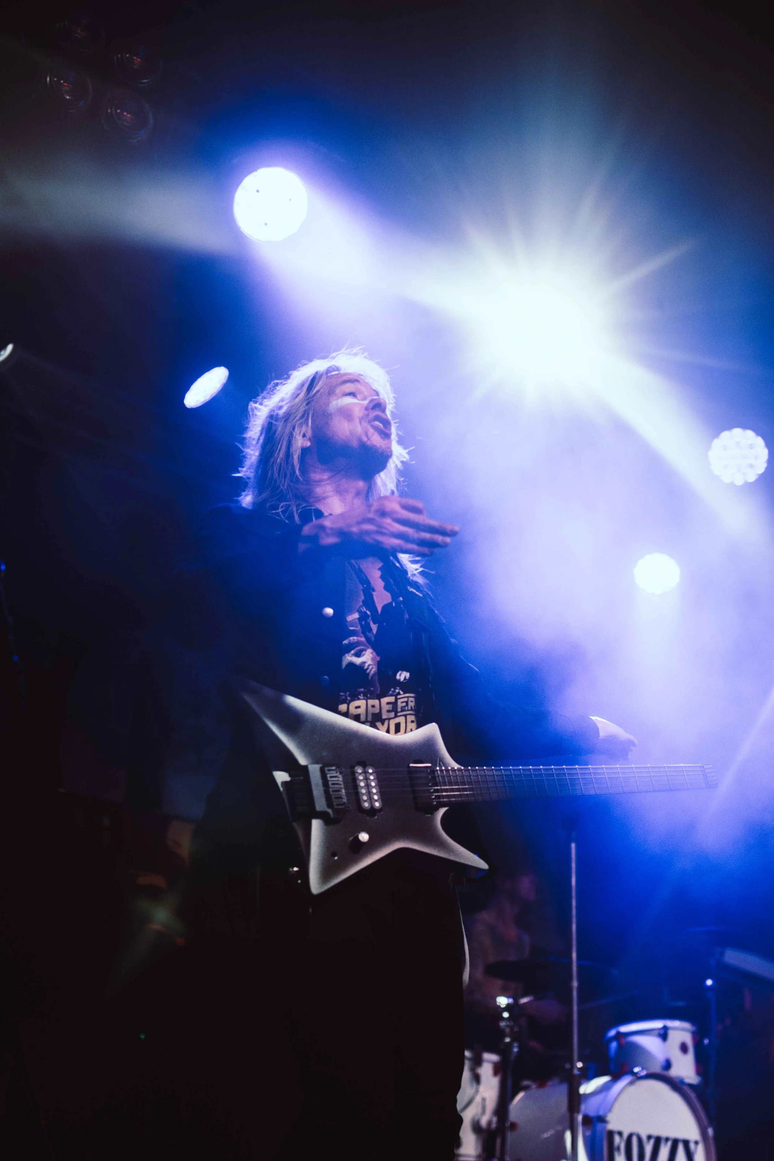 A musician with long blonde hair playing an electric guitar on stage, illuminated by bright stage lights, with drums in the background.