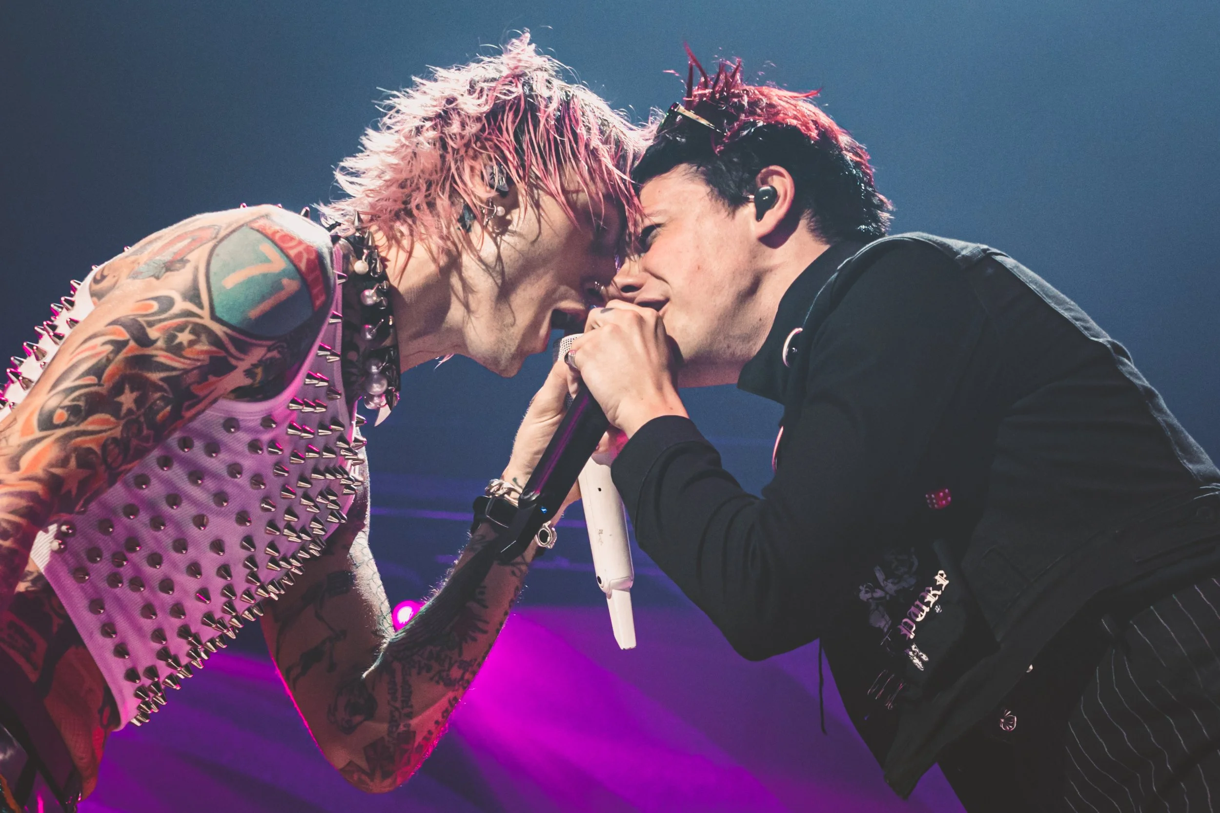 Two male performers with pink and red hair, tattoos, and emo-style clothing are singing into a microphone on stage with purple lighting.